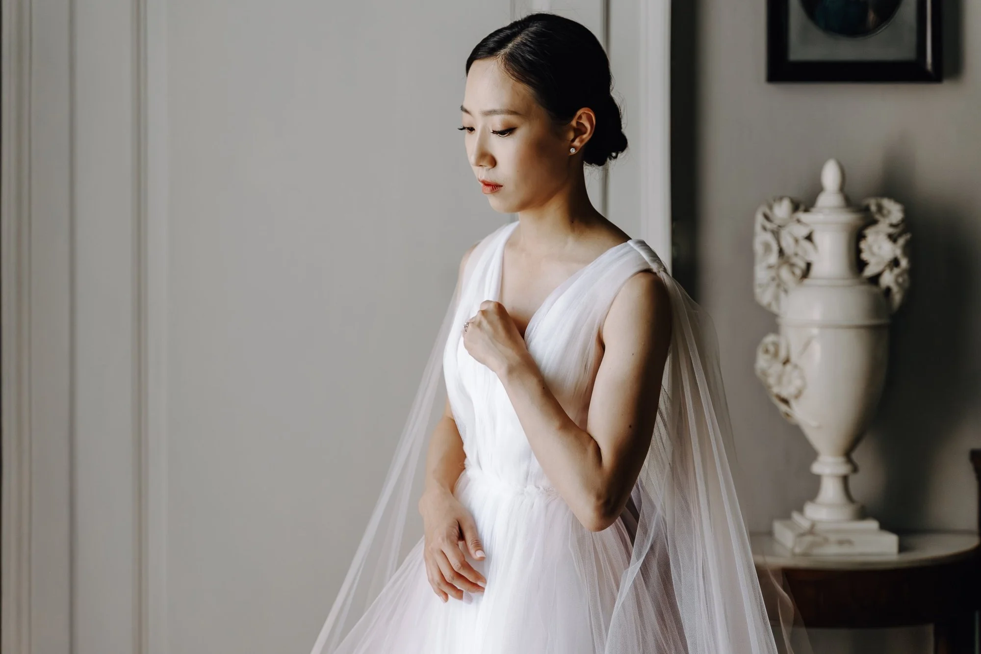A woman in a white dress looking down and holding her chest, standing next to a decorative vase on a table in a room with minimalistic decor.