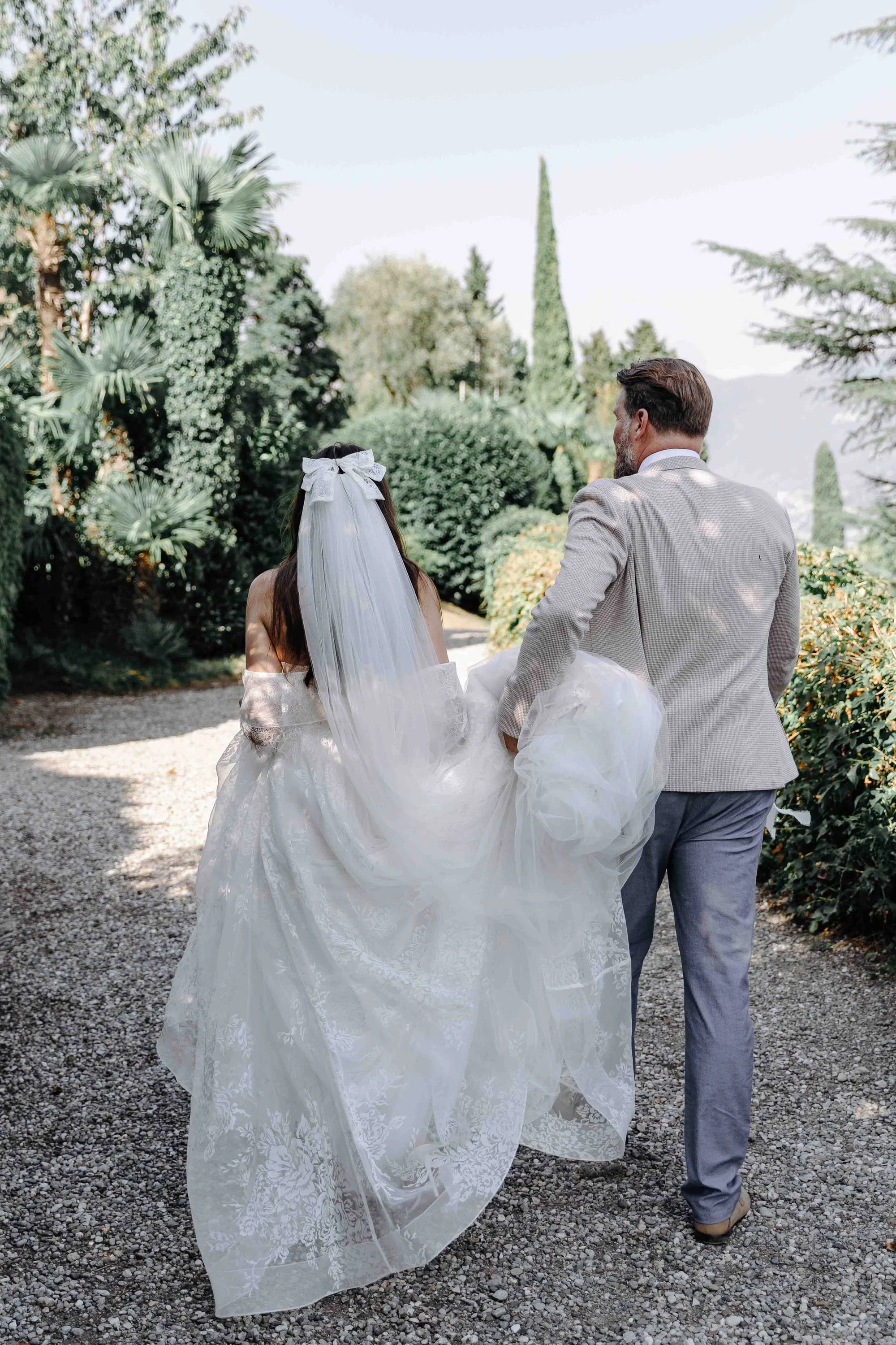 A bride and groom walking outdoors on a gravel path surrounded by greenery, with the bride wearing a white wedding dress and veil, and the groom in a light gray suit.