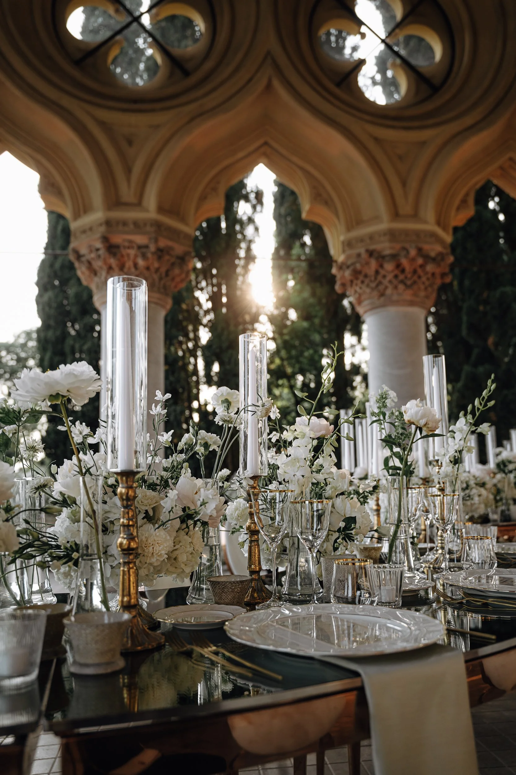 Elegant outdoor dining table decorated with white flowers and tall glass candle holders, set under a decorative archway with intricate columns and a sunset in the background.
