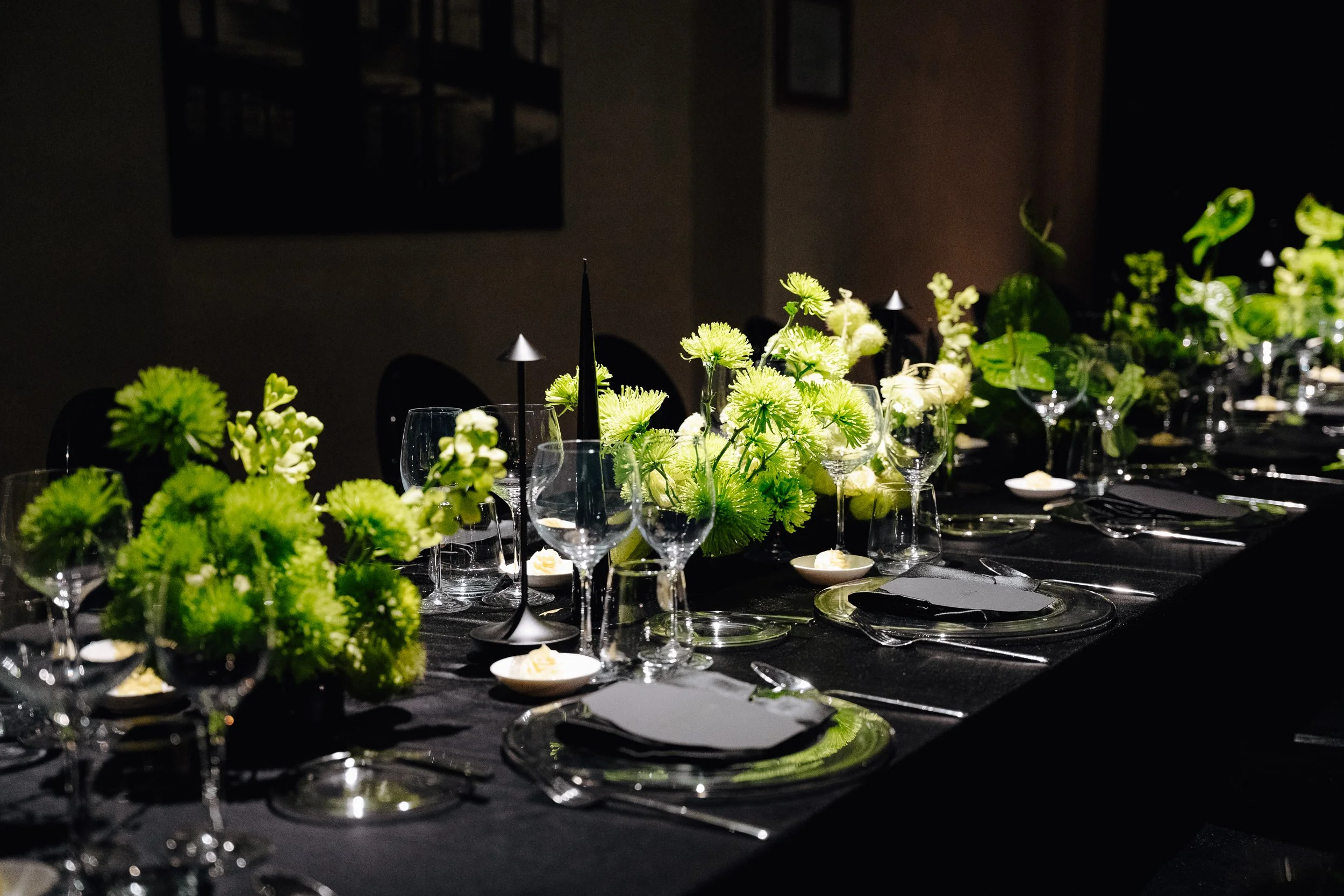 A round dining table decorated with a black tablecloth, featuring a centerpiece of green and white flowers, along with wine glasses, small plates, and black napkins.