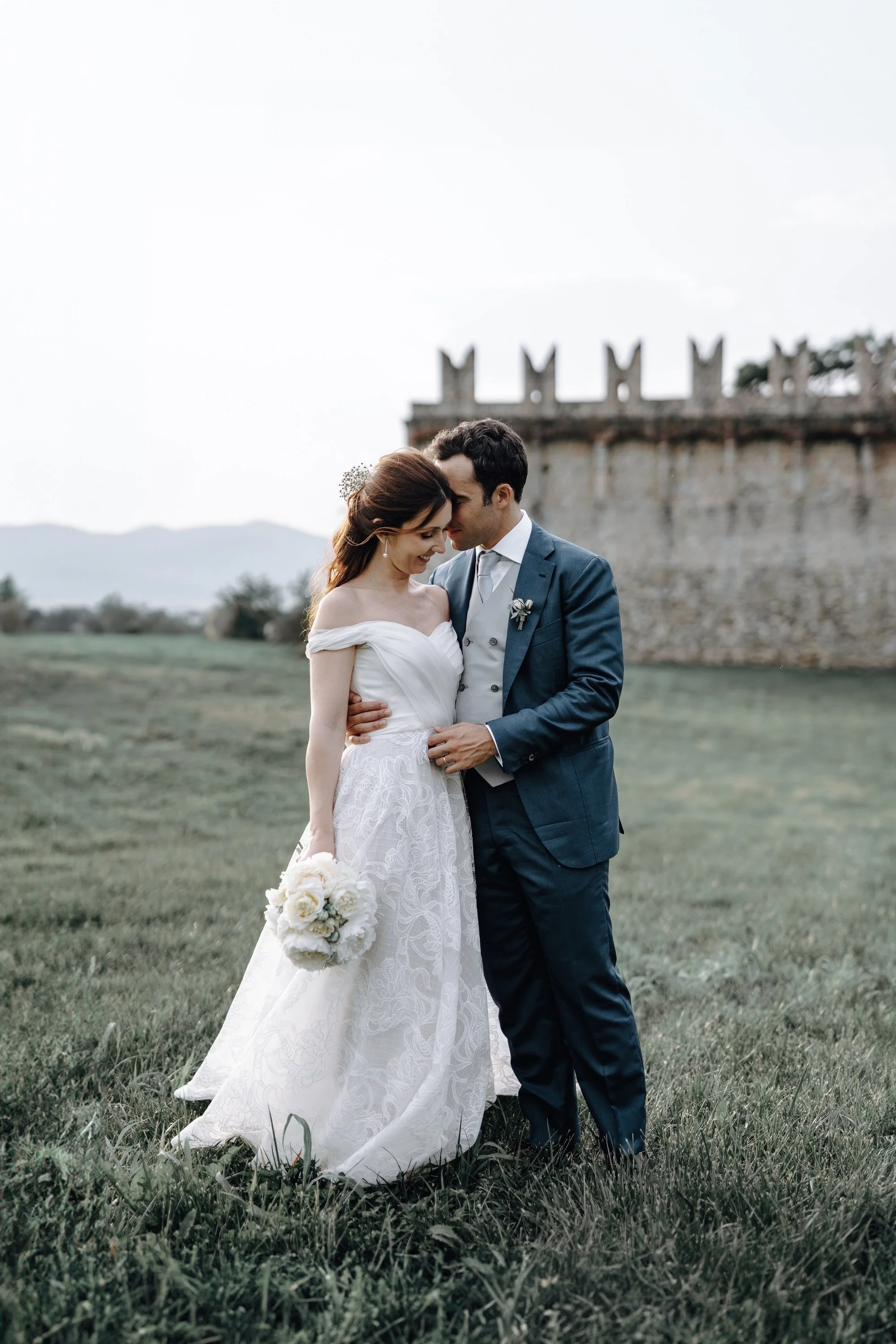 A newlywed couple stands close together in a grassy field with an historic castle in the background. The bride wears a white off-the-shoulder wedding dress and holds a bouquet of white flowers, while the groom wears a dark blue suit and white shirt. They are touching foreheads and smiling.