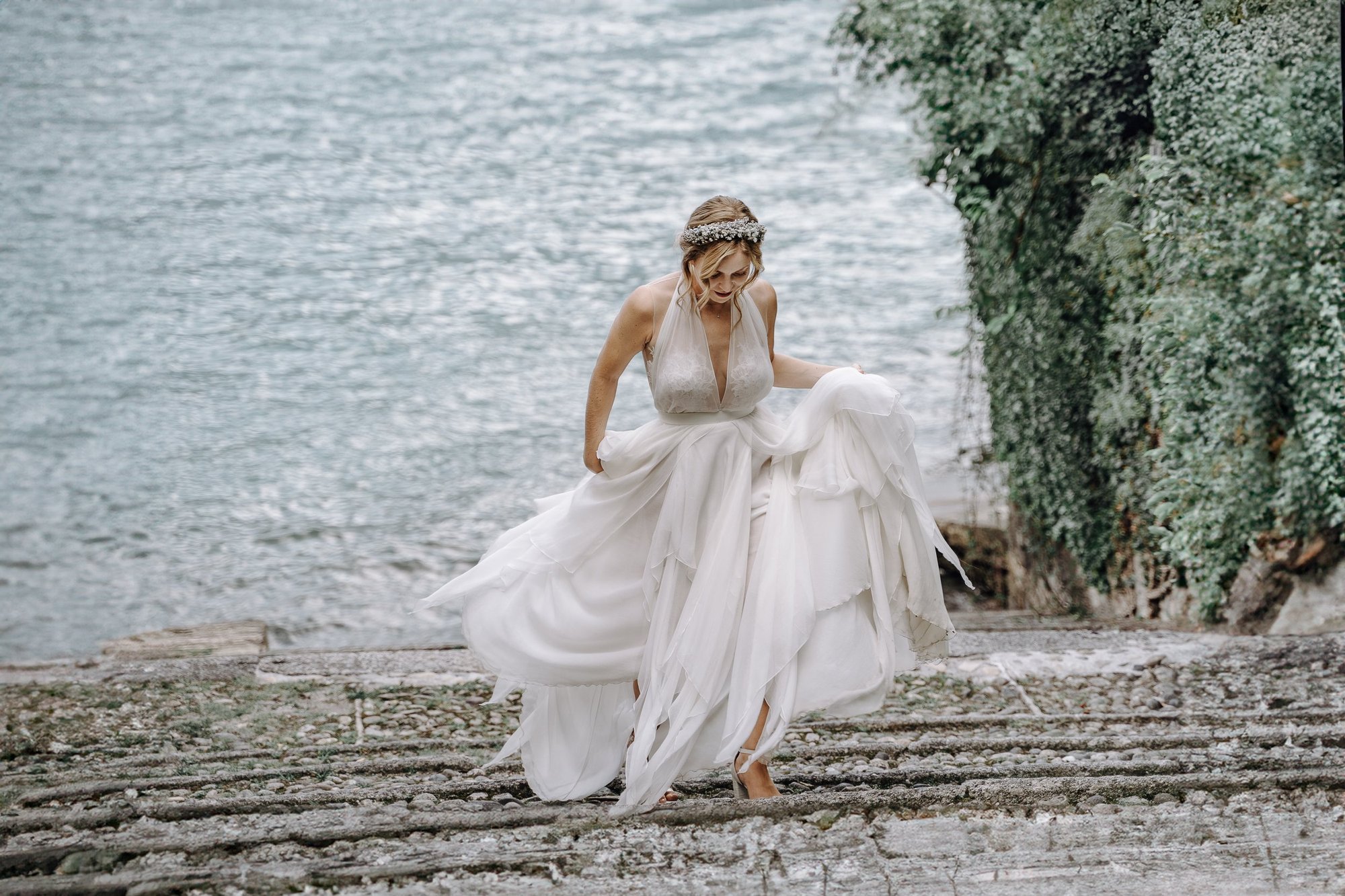 A woman in a white wedding dress standing on a rocky surface near a body of water, with greenery on the right side, holding her dress up slightly and looking down.