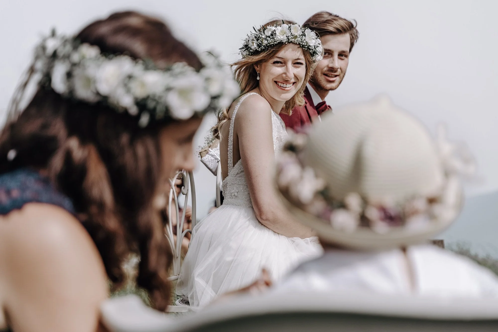 A woman in a wedding dress and floral crown, smiling, sitting at what appears to be a wedding or celebration, with other guests around her.