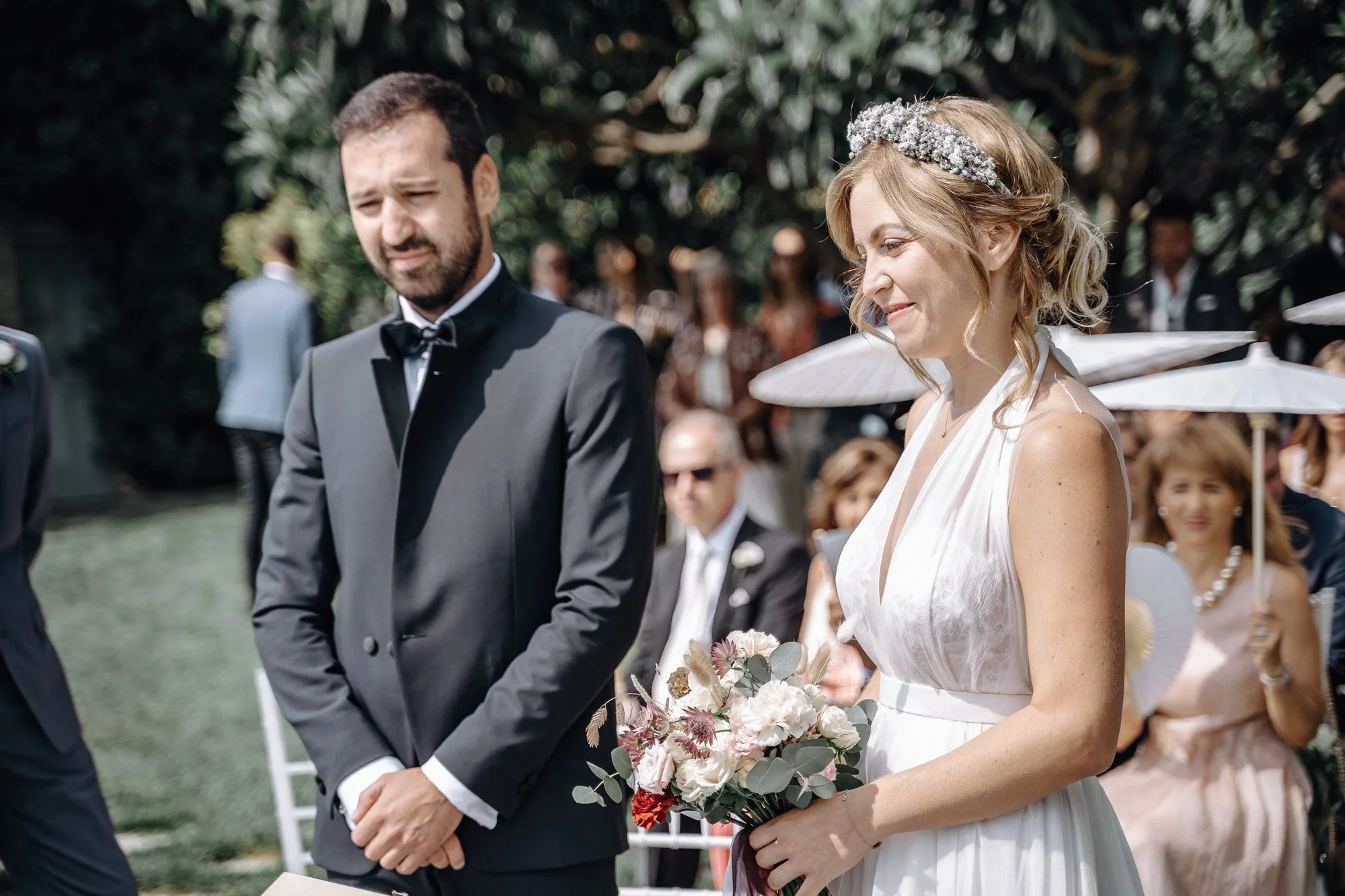 A bride and groom standing together during a wedding ceremony outdoors. The bride is smiling and holding a bouquet of white and light-colored flowers, while the groom appears solemn with eyes closed. The background shows guests seated under white umb