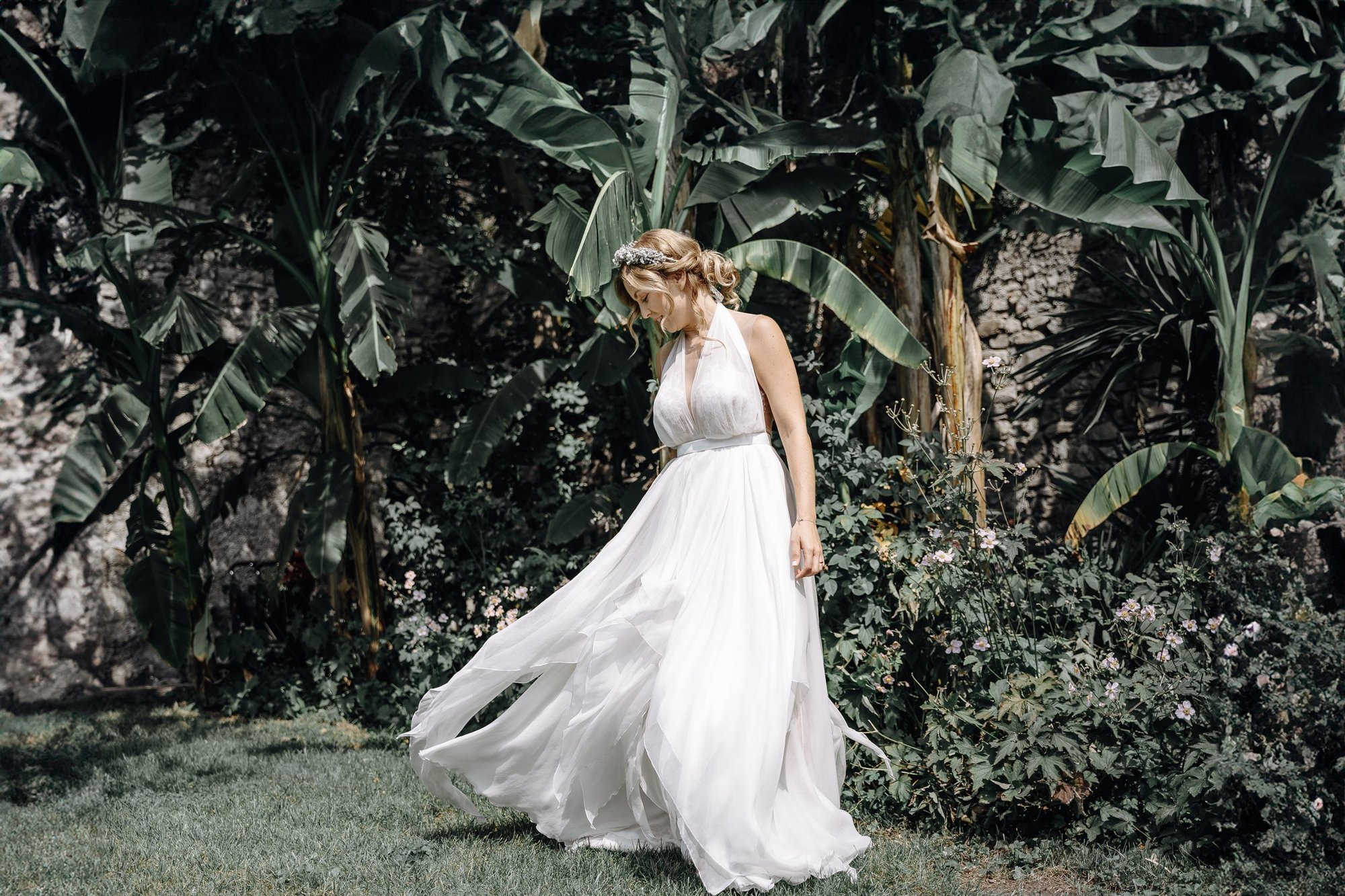 A woman in a white dress standing outdoors among large green tropical leaves and flowering bushes, looking down, with her hair styled in loose curls and wearing a floral headband.