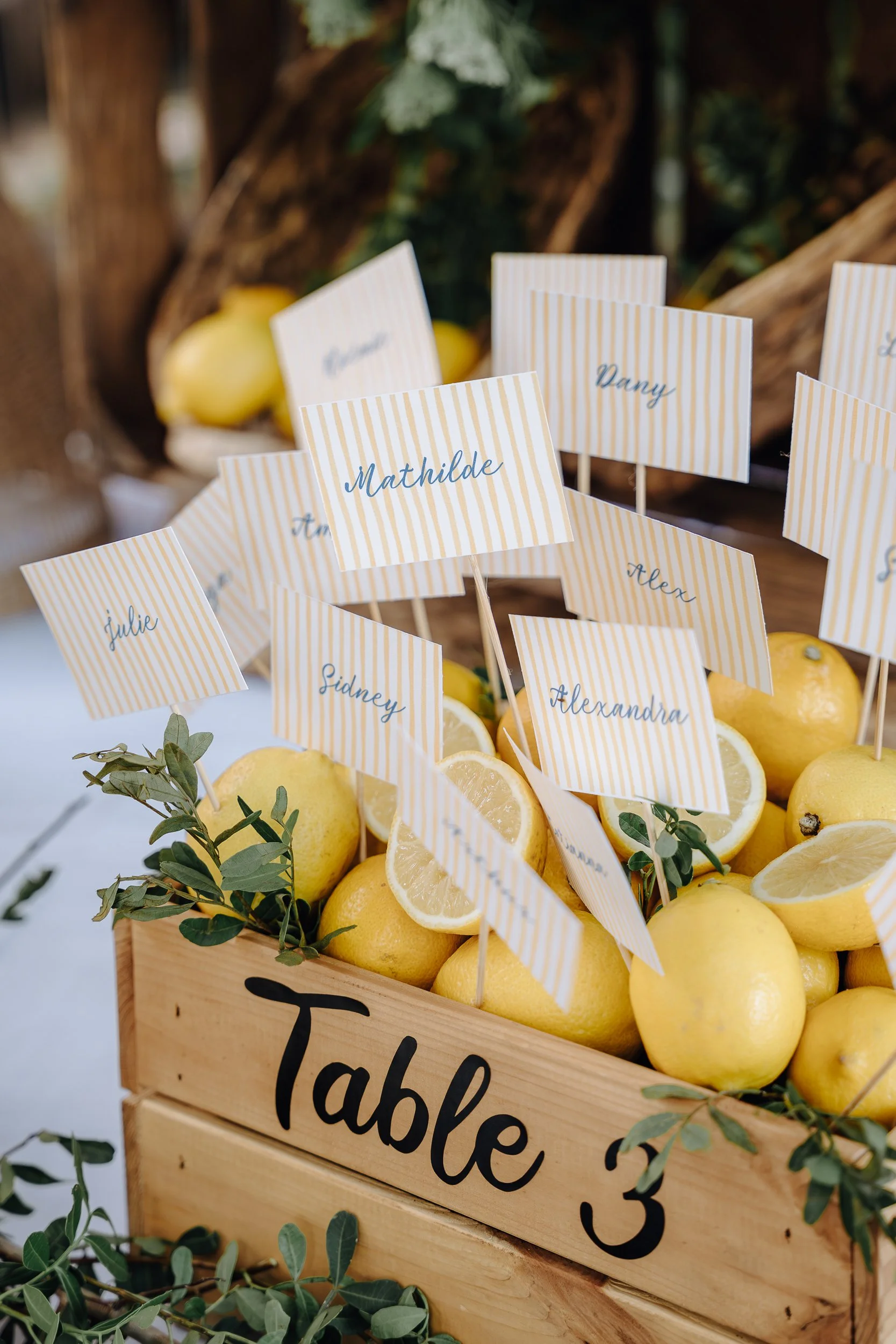A wooden table labeled 'table 3' with a basket of lemons, some sliced and some whole, decorated with small green leaves, and place cards with handwritten names and stripes in the background.