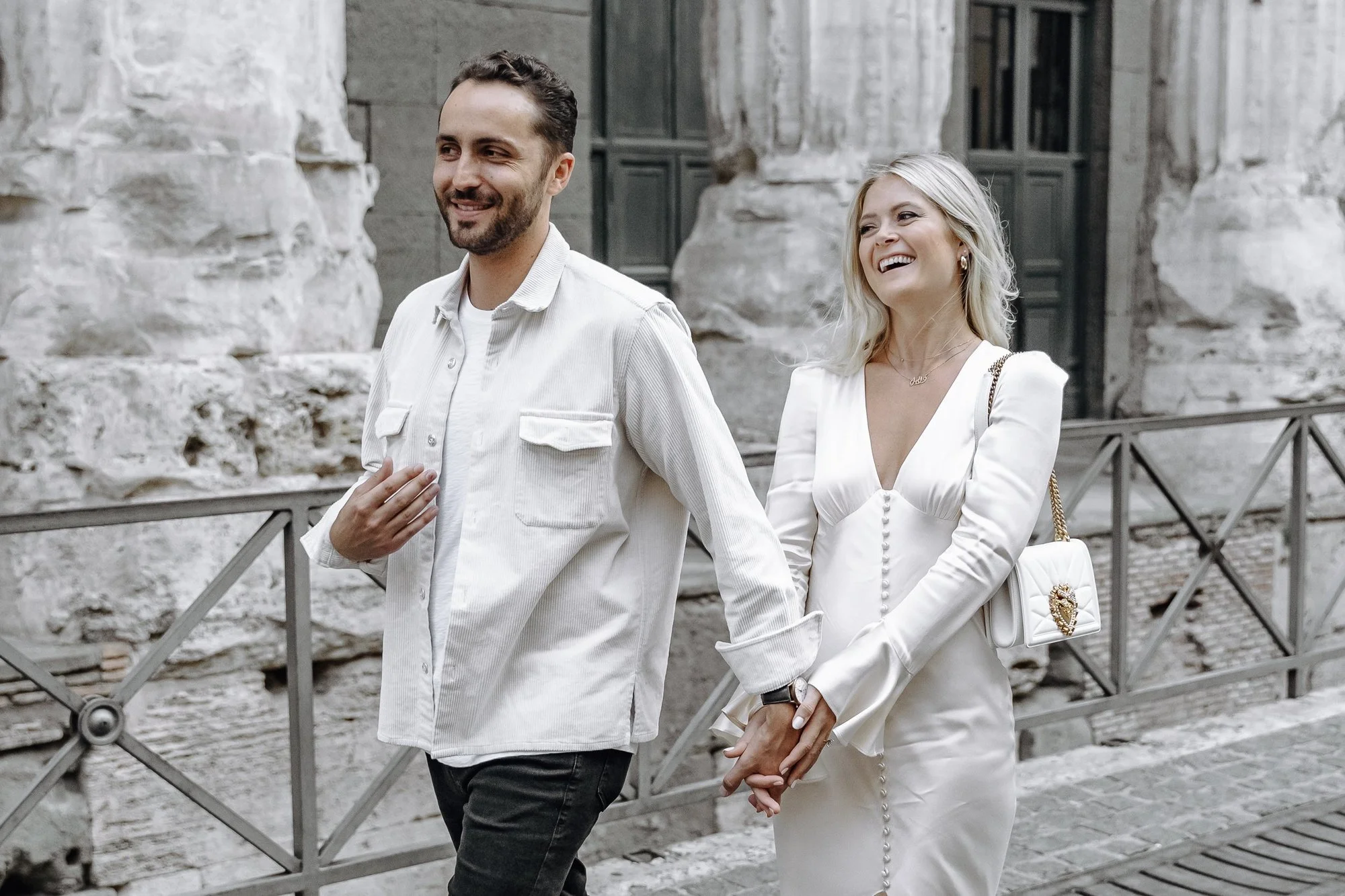 A smiling man and woman walking hand in hand in front of an ancient stone building with large dark windows. The man has a beard and is wearing a white shirt, and the woman has blonde hair and is wearing a white dress and carrying a white handbag.