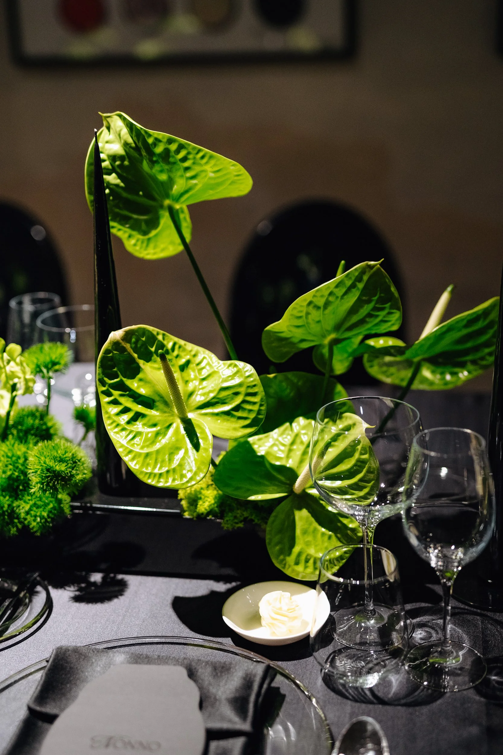 A close-up view of a floral arrangement featuring green anthurium and other plants on a dark table, with clear wine glasses and a small dish of butter in the foreground.