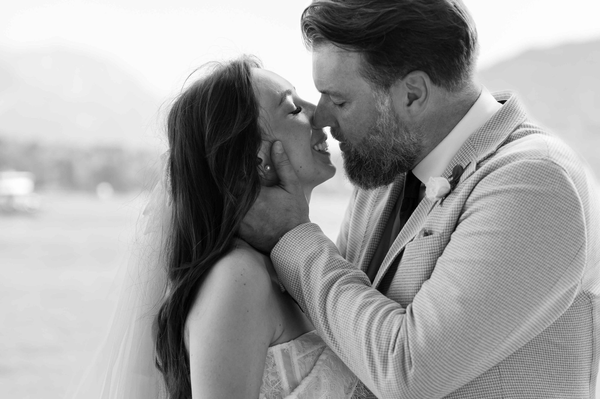 A black and white photo of a couple in wedding attire, close together, about to kiss outdoors.