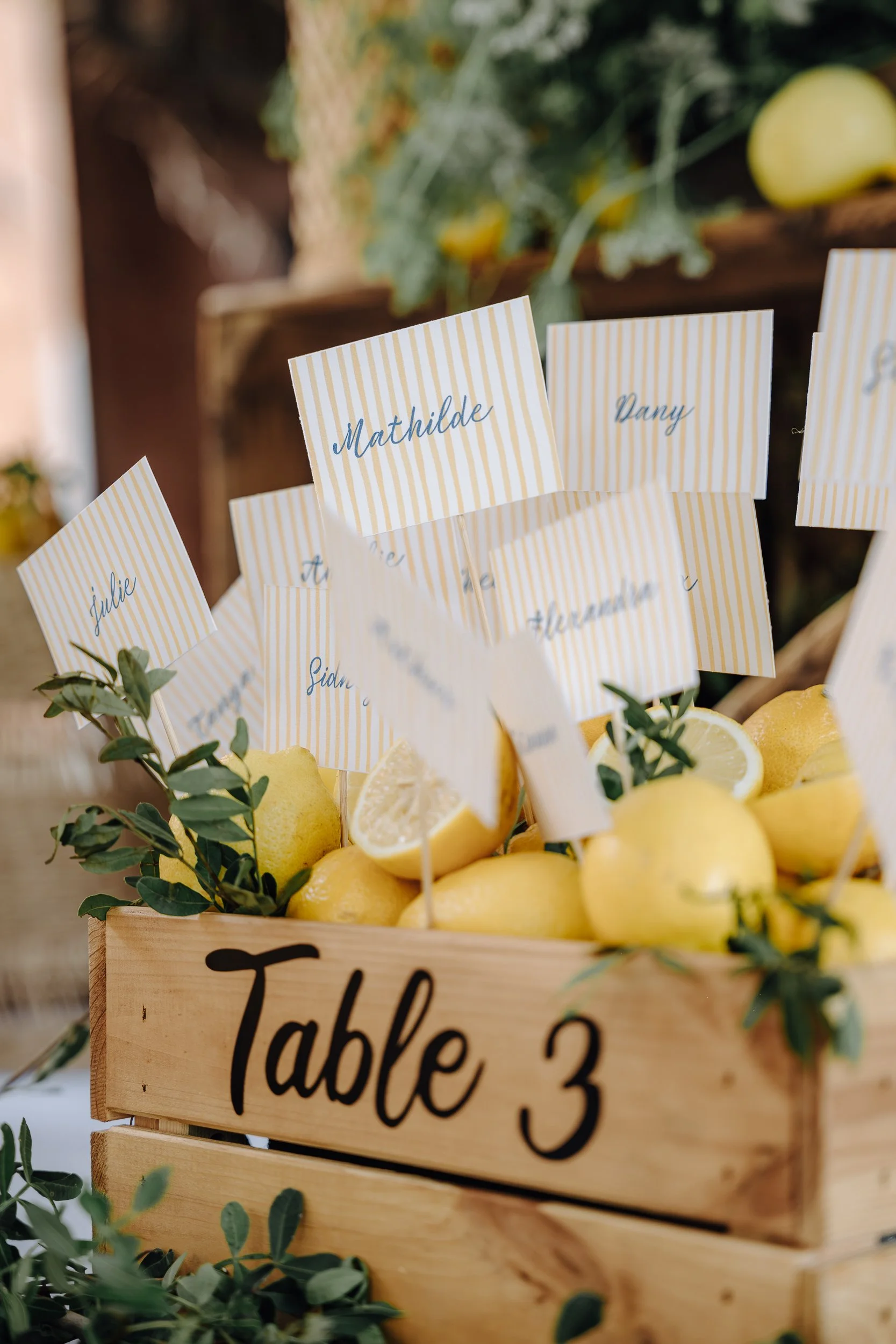 A wooden table sign with black cursive letters reading 'Table 3' surrounded by lemons, lemon slices, and green leaves, with small paper flags with striped yellow and white backgrounds and handwritten names attached to toothpicks, placed among the lem