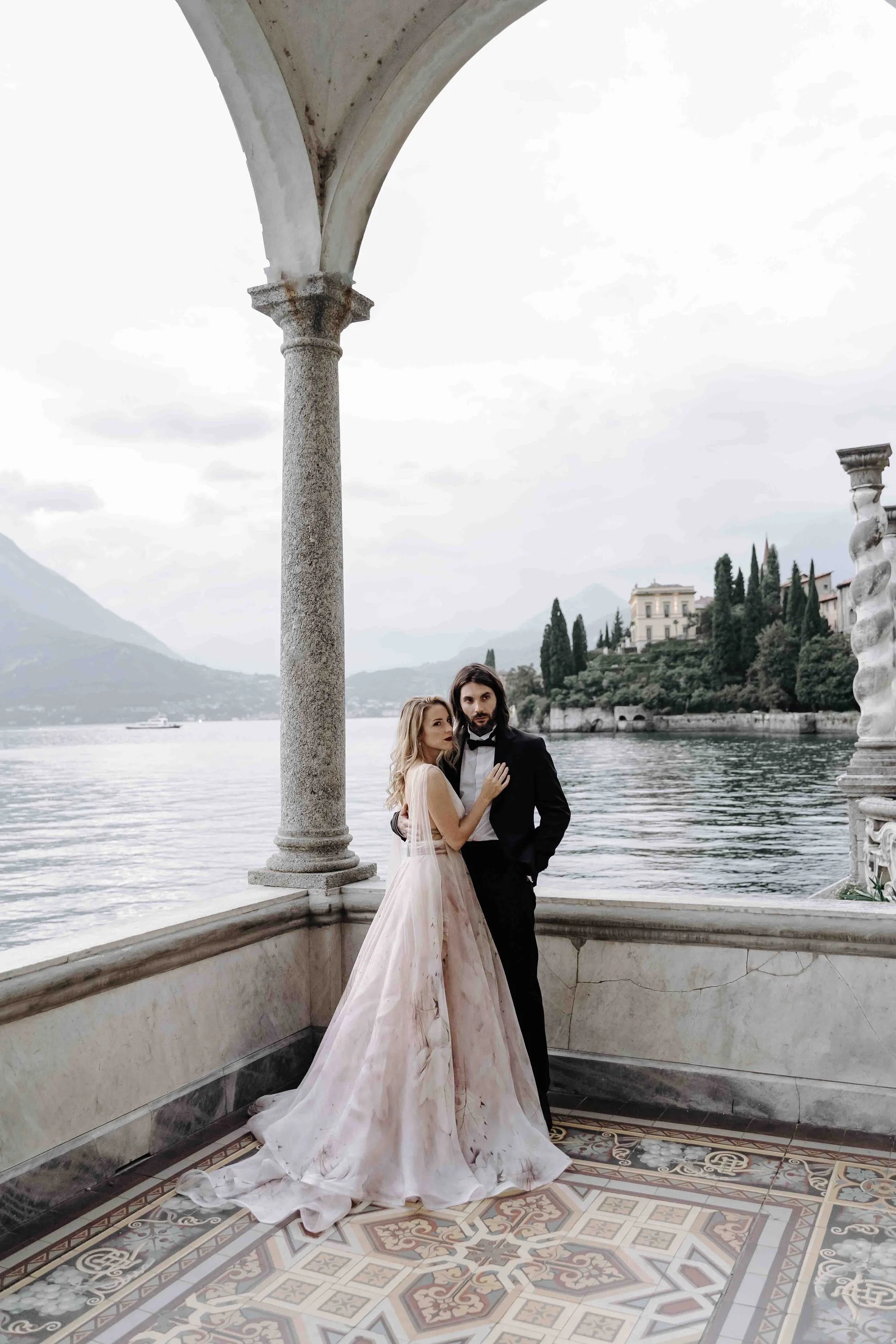 A couple dressed in formal attire as a bride and groom, standing on a balcony by the water with scenic hills and buildings in the background. The bride is wearing an elegant pink and white gown, and the groom is in a black tuxedo.
