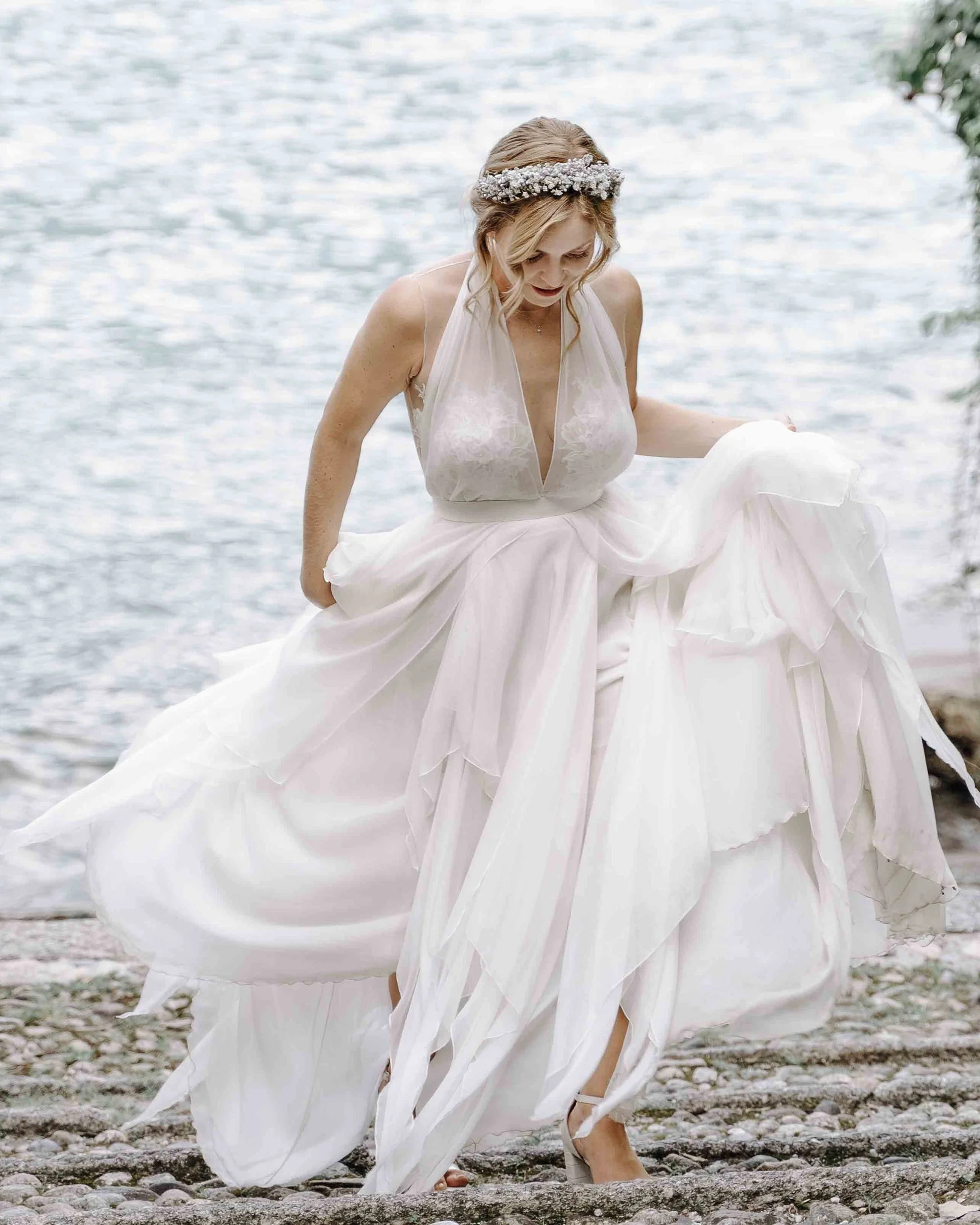 A woman in a wedding dress is standing outdoors near a body of water, holding up her dress with one hand and wearing high heels and a floral headband.