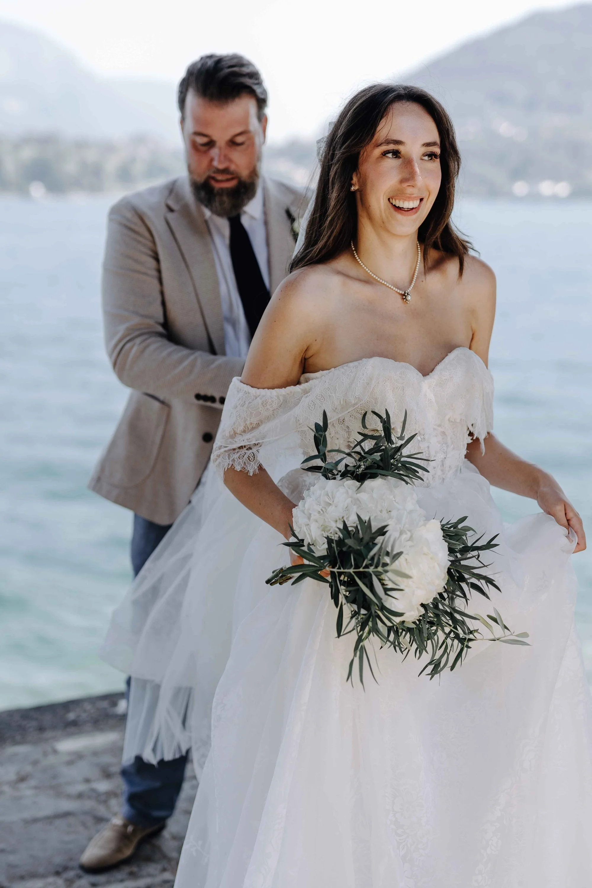 A bride in a white off-the-shoulder wedding dress holding a bouquet of white flowers and greenery, smiling, with a groom in a tan suit and dark tie standing behind her, near a body of water with a mountain and cloudy sky in the background.