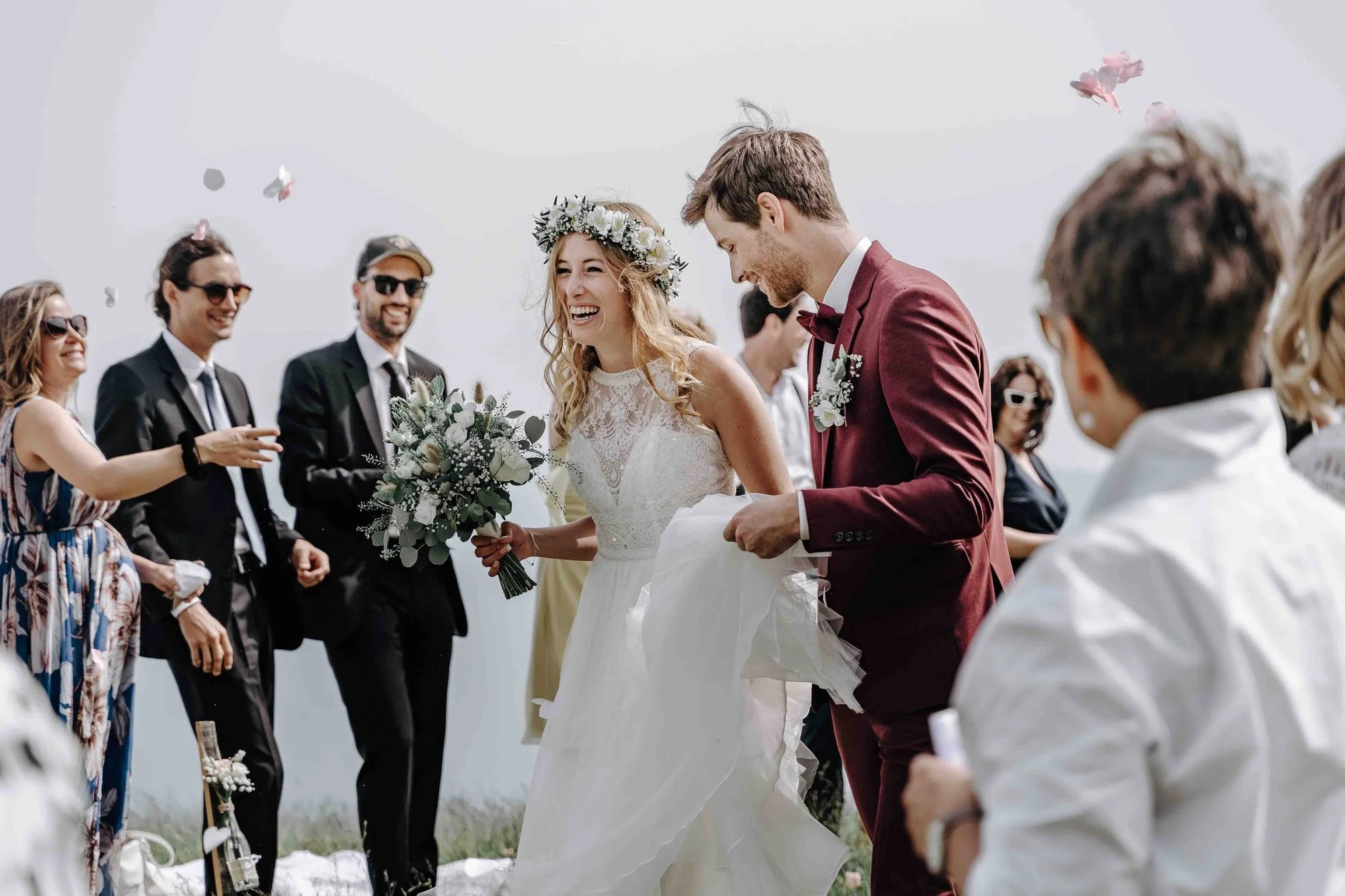 A bride and groom share a moment of happiness during their wedding ceremony outdoor with friends and family in the background, some wearing sunglasses and smiling, with pink flower petals falling around them.
