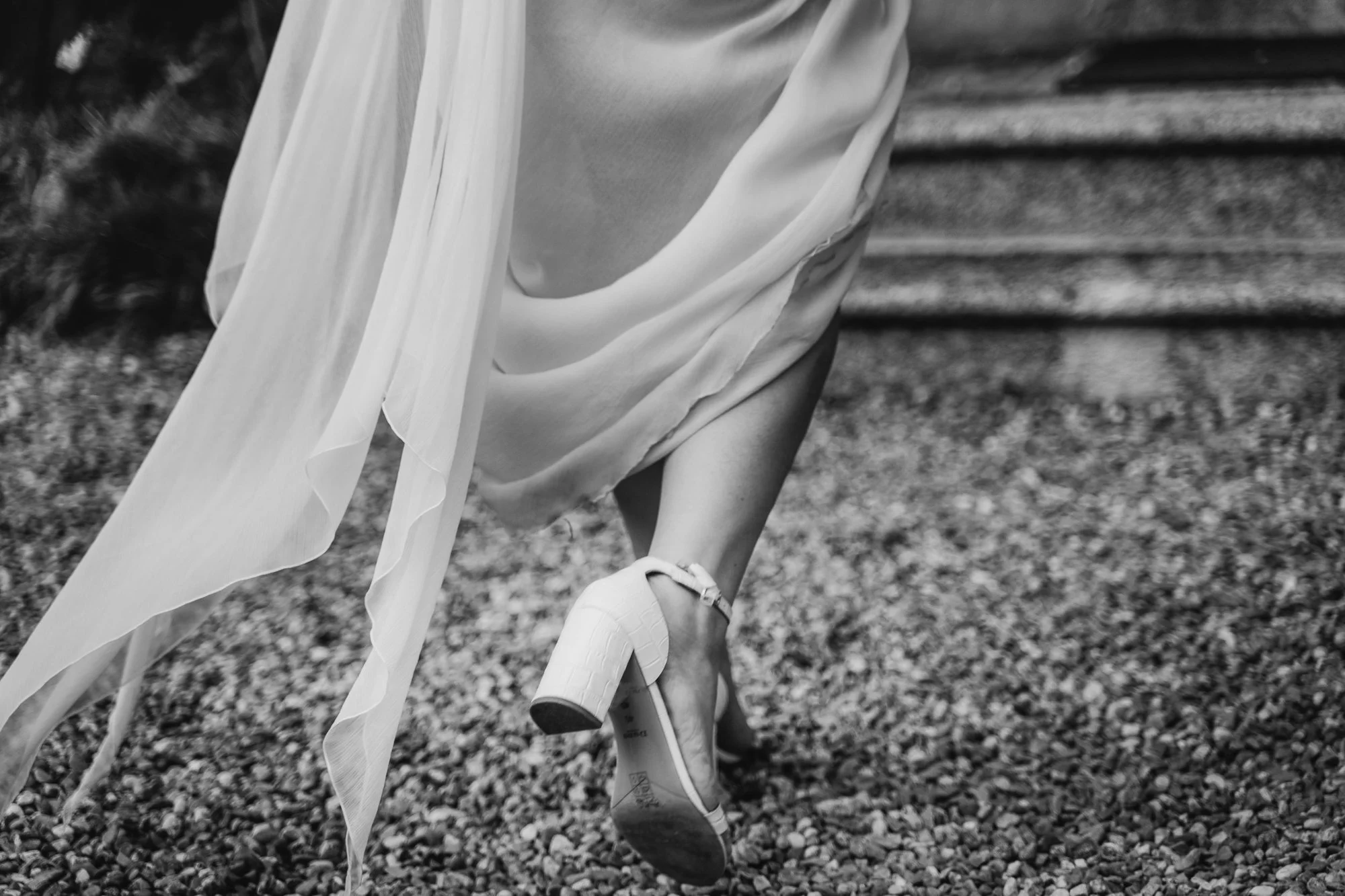 A person dressed in a white flowing dress with high heels, walking on gravel outdoors, with steps visible in the background.