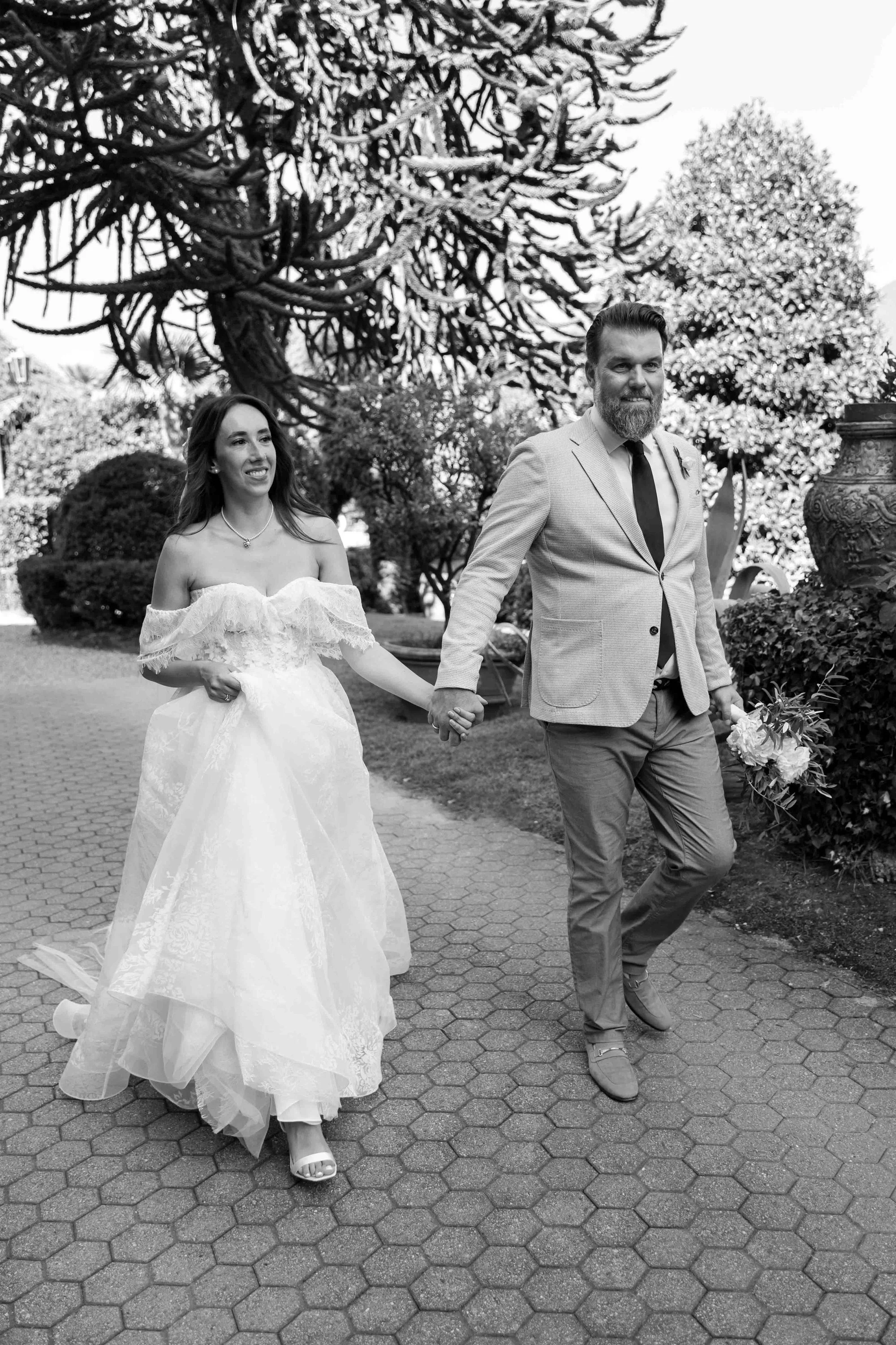 A black-and-white photograph of a bride and groom walking hand-in-hand outdoors. The bride is wearing a strapless wedding gown with lace details and holding part of her dress with her left hand. The groom is wearing a light-colored suit with a tie an