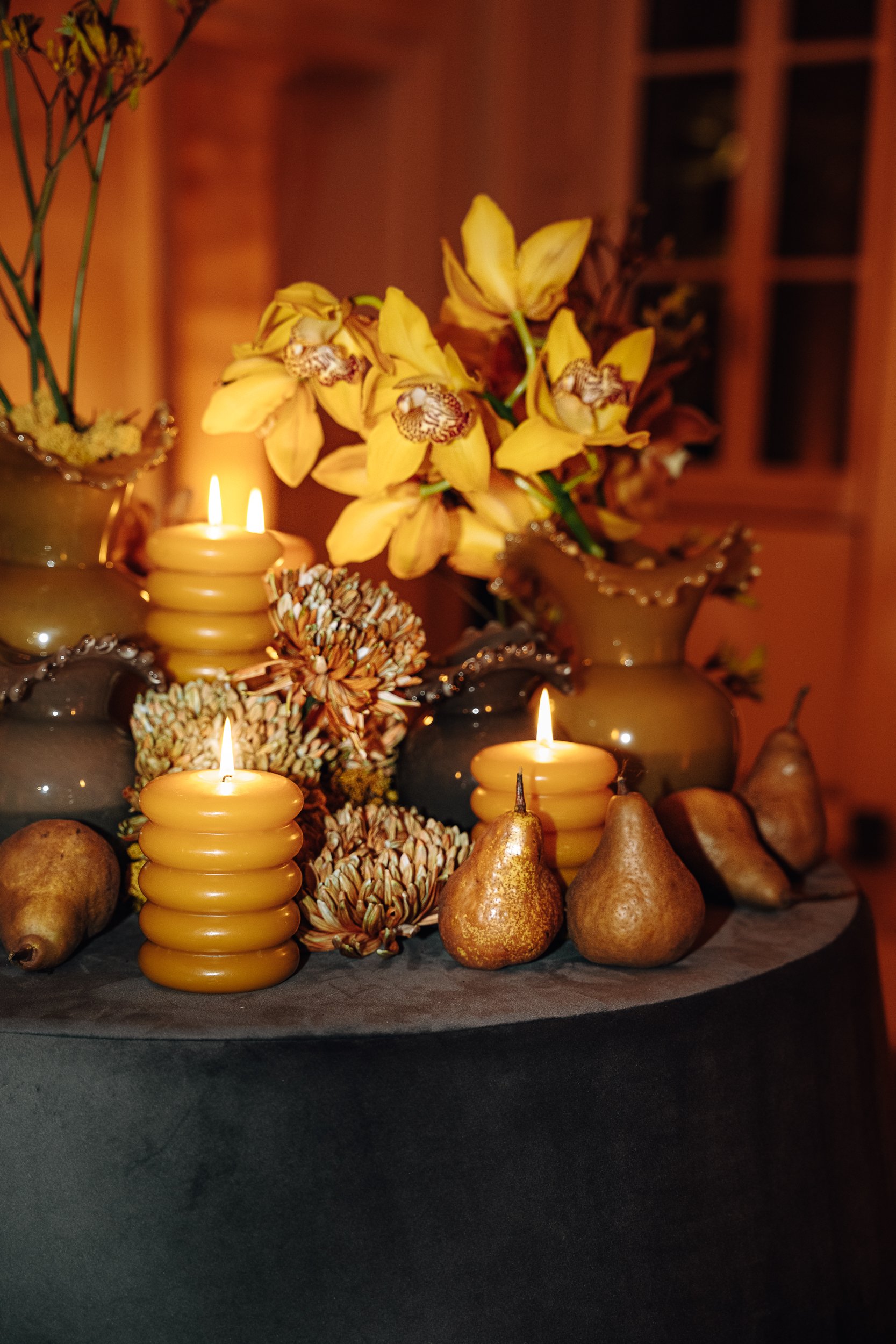 A fall centerpiece with yellow orchids, candles, pears, and decorative pinecones arranged on a black tablecloth.