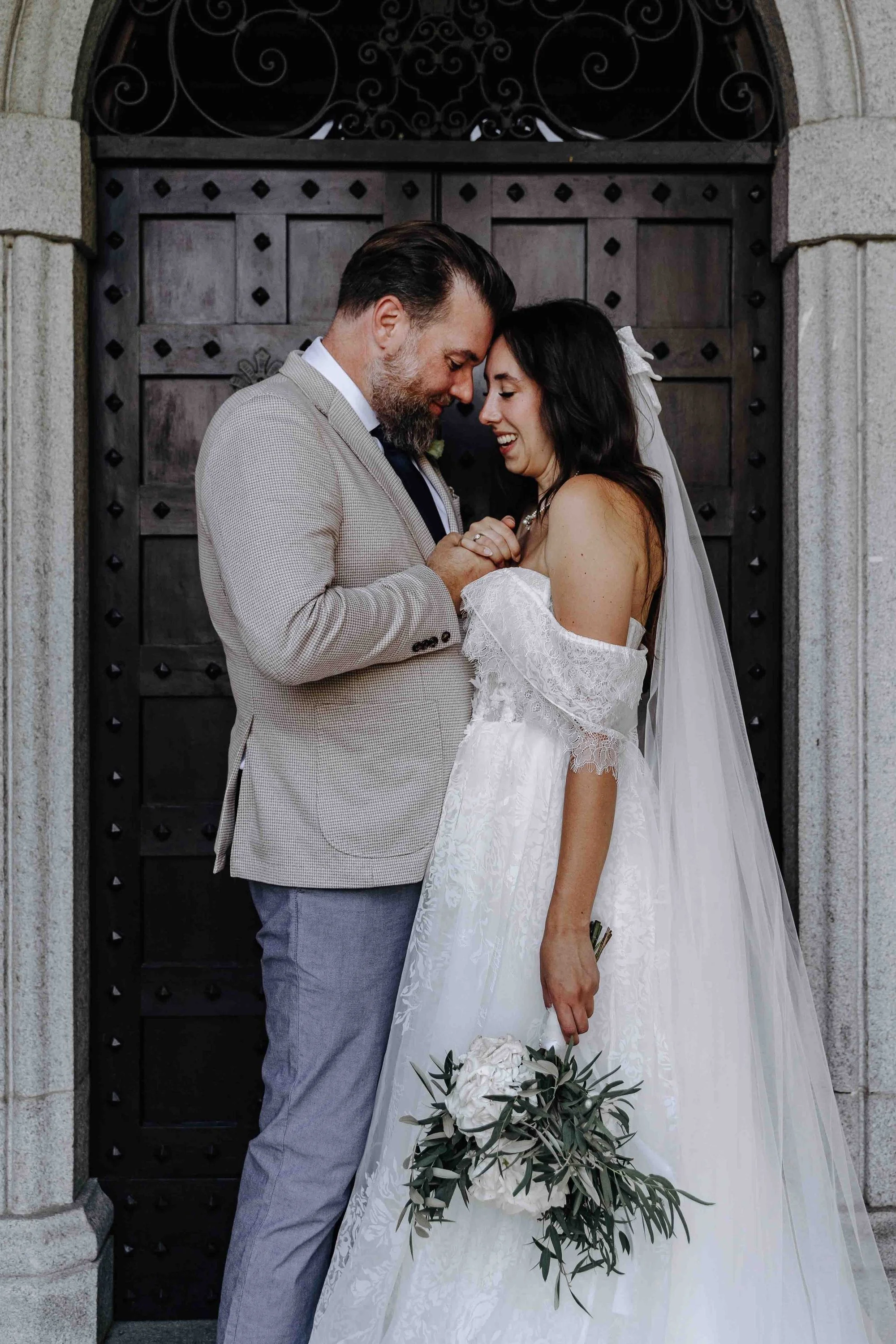 A bride and groom stand very close, holding hands and smiling with their foreheads touching in front of a large wooden door with black metal accents. The bride wears a white wedding dress with off-the-shoulder lace and has a long veil, while the groo