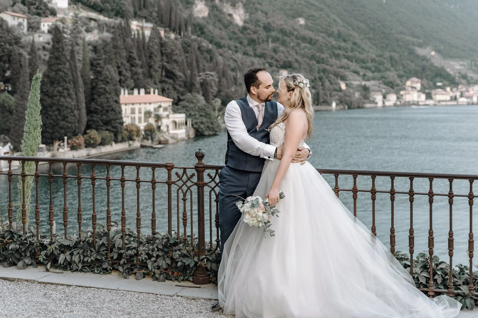 A newlywed couple shares a romantic moment on a lakeside terrace with a scenic mountain backdrop. The bride wears a white wedding gown and holds a bouquet, while the groom is dressed in a dark vest and light shirt. They are embracing as they lean int
