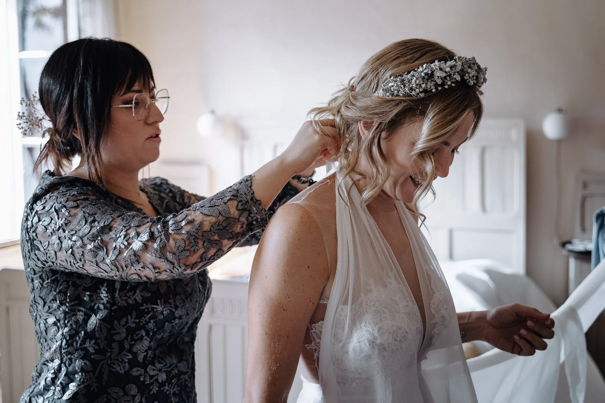 A woman with blonde hair wearing a lace dress is having her hair arranged by another woman in a patterned dress, who appears to be helping her get ready, likely for a special occasion or wedding.