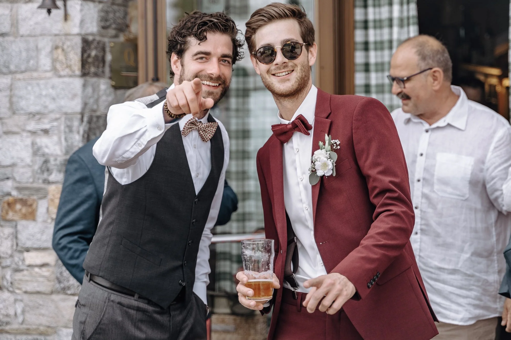 Two men are smiling at a gathering, with one pointing at the camera. The man on the left has curly hair and wears a vest and bow tie, while the man on the right has straight hair, sunglasses, a red suit, and a boutonniere. A third man in glasses and 