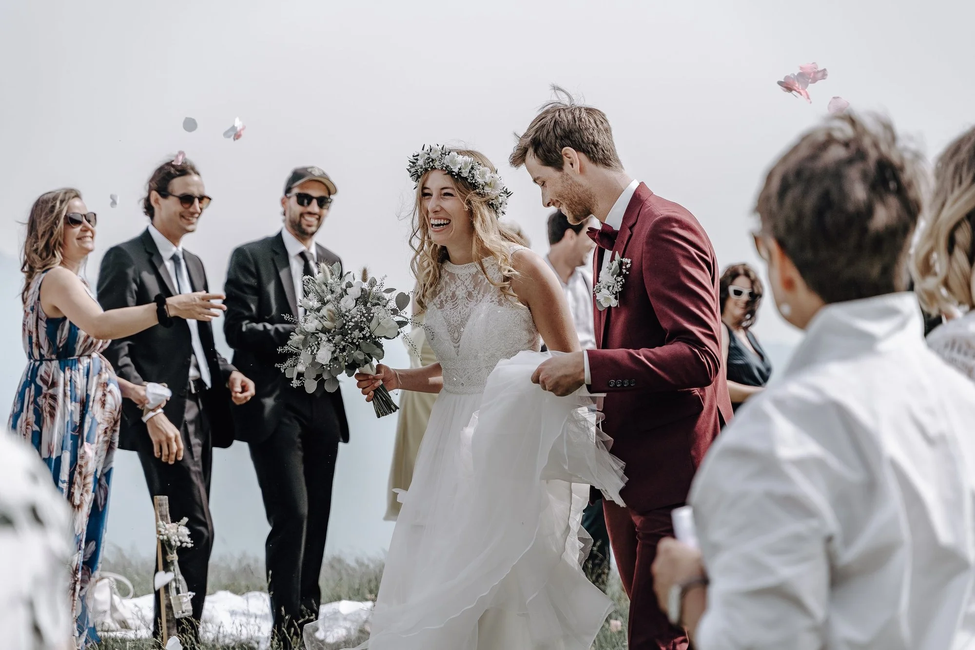 A wedding ceremony with a bride and groom, surrounded by guests, outdoors with a cloudy sky. The bride is holding a bouquet and wearing a lace dress with a floral crown. The groom is dressed in a maroon suit with a bow tie. Guests are smiling, wearin