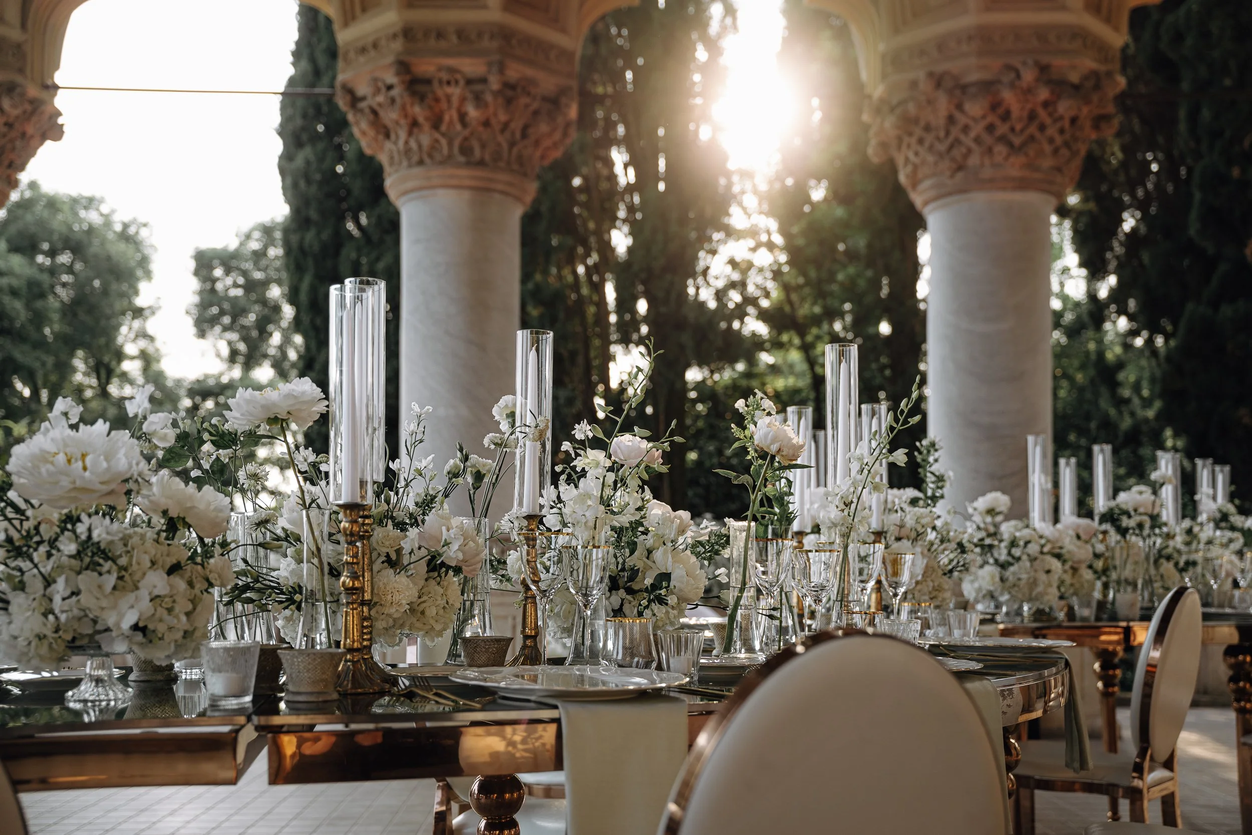 Elegant outdoor dining table decorated with white flowers, tall glass candle holders, and gold accents, set in front of classical columns with ornate capitals, sun shining through trees in the background.