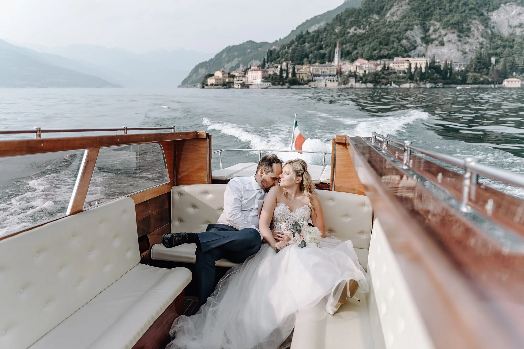 A couple dressed in wedding attire sitting together on a boat, with a scenic lake and hillside town in the background.