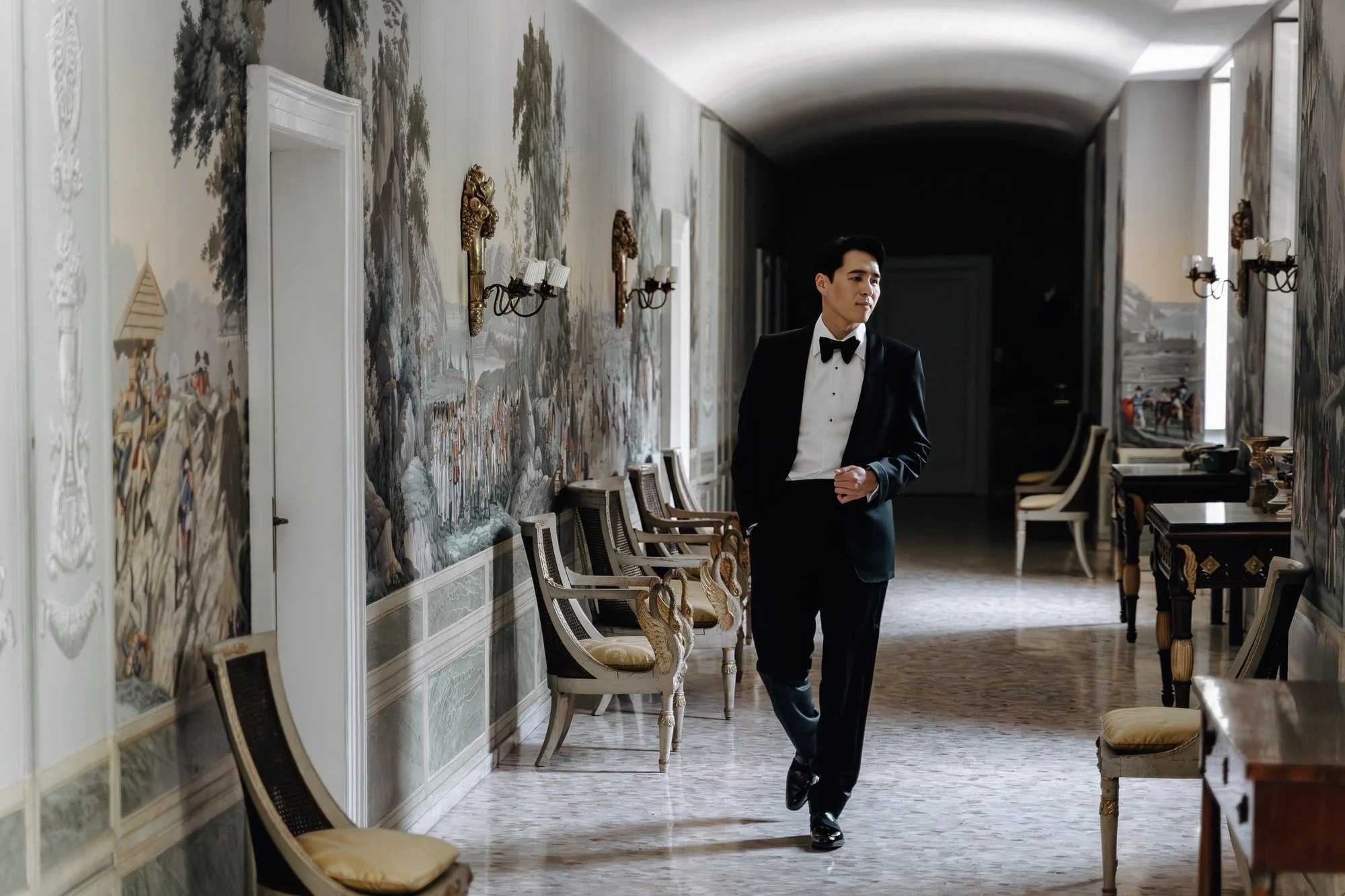 A man in a tuxedo walking through an elegant, ornate room with patterned wallpaper, antique chairs, and large windows.