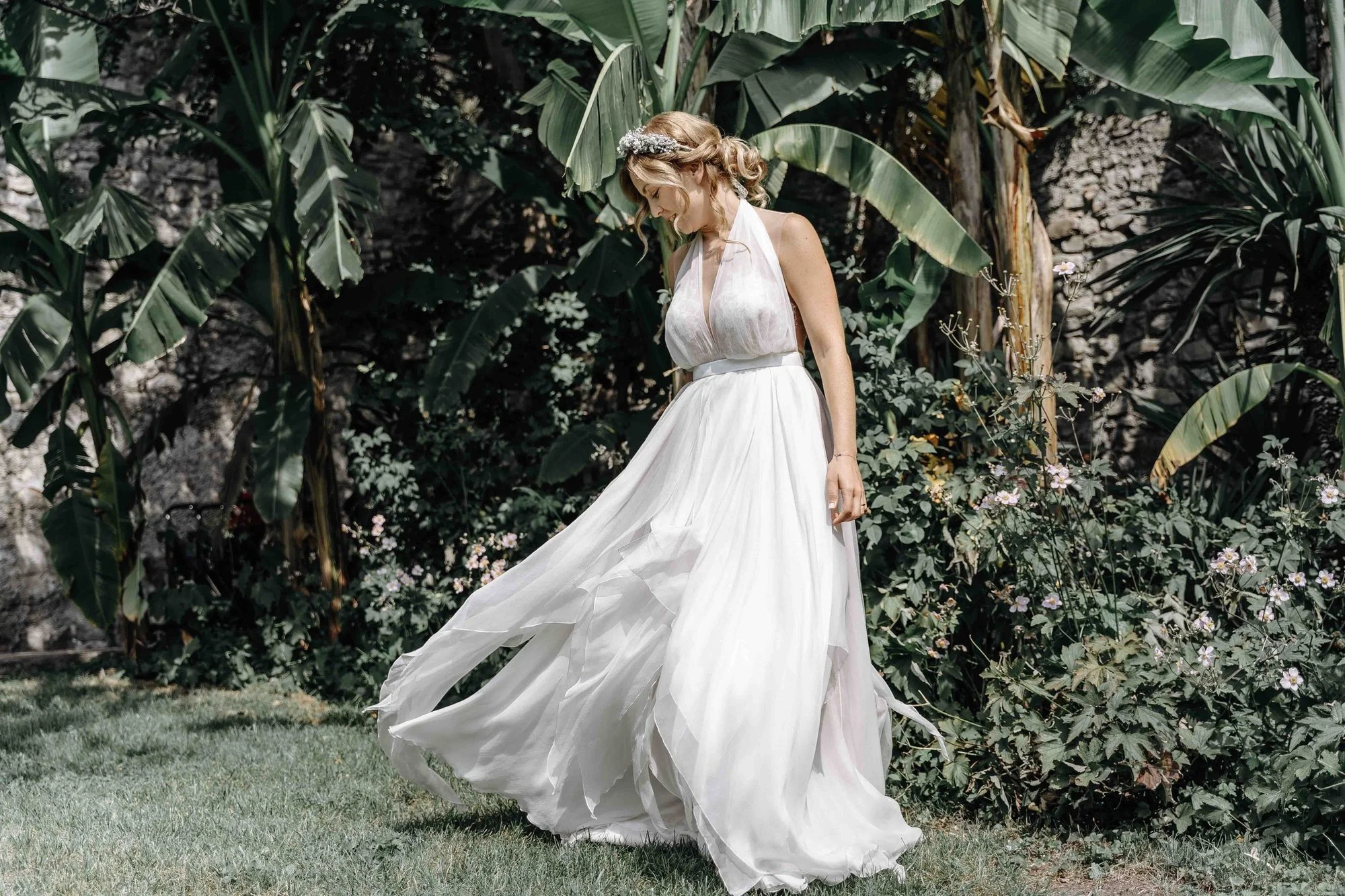 A woman in a flowing white dress standing among lush green banana plants and flowering bushes.