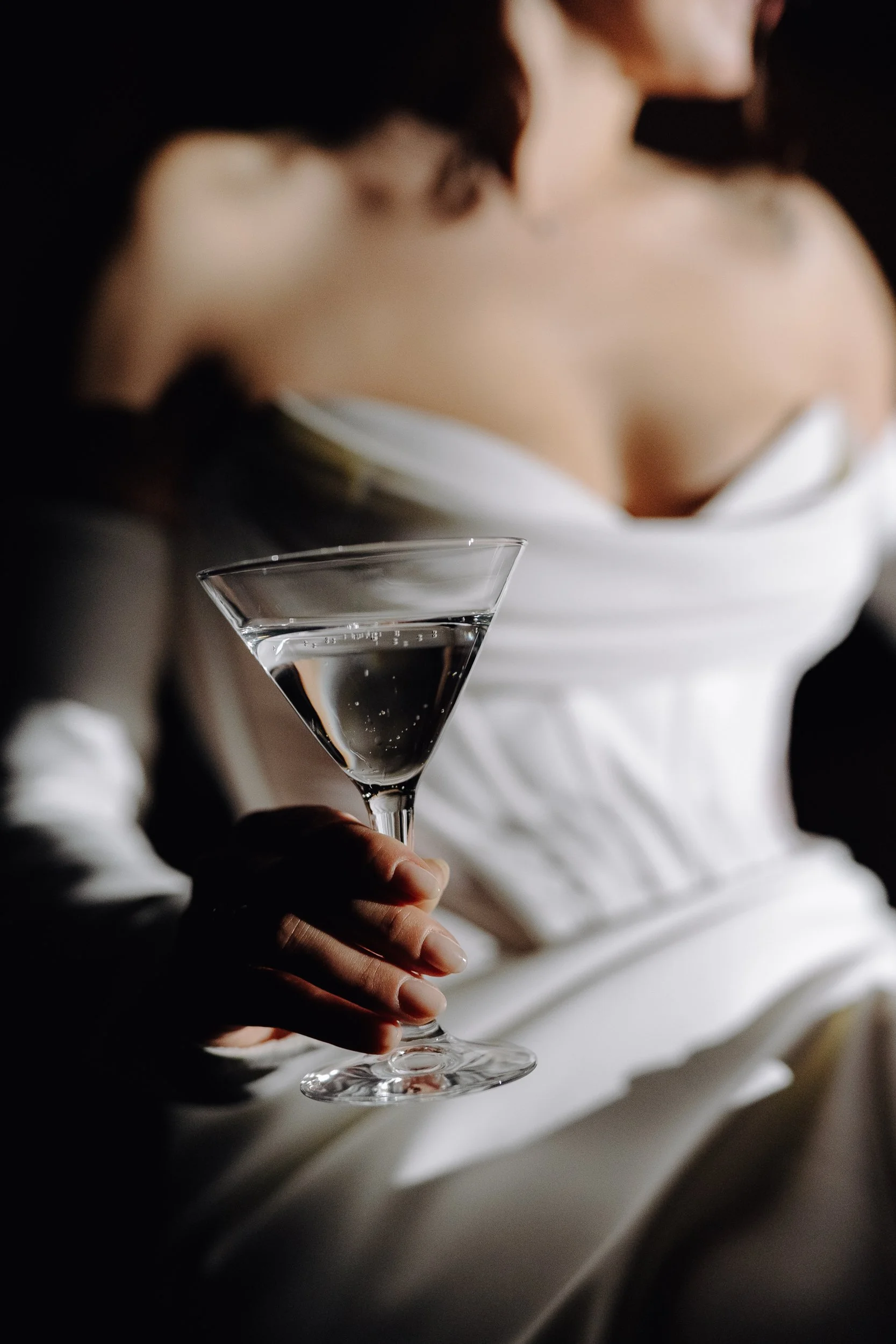 A person holding a martini glass filled with a clear liquid, with a blurred background of a woman wearing a white strapless dress