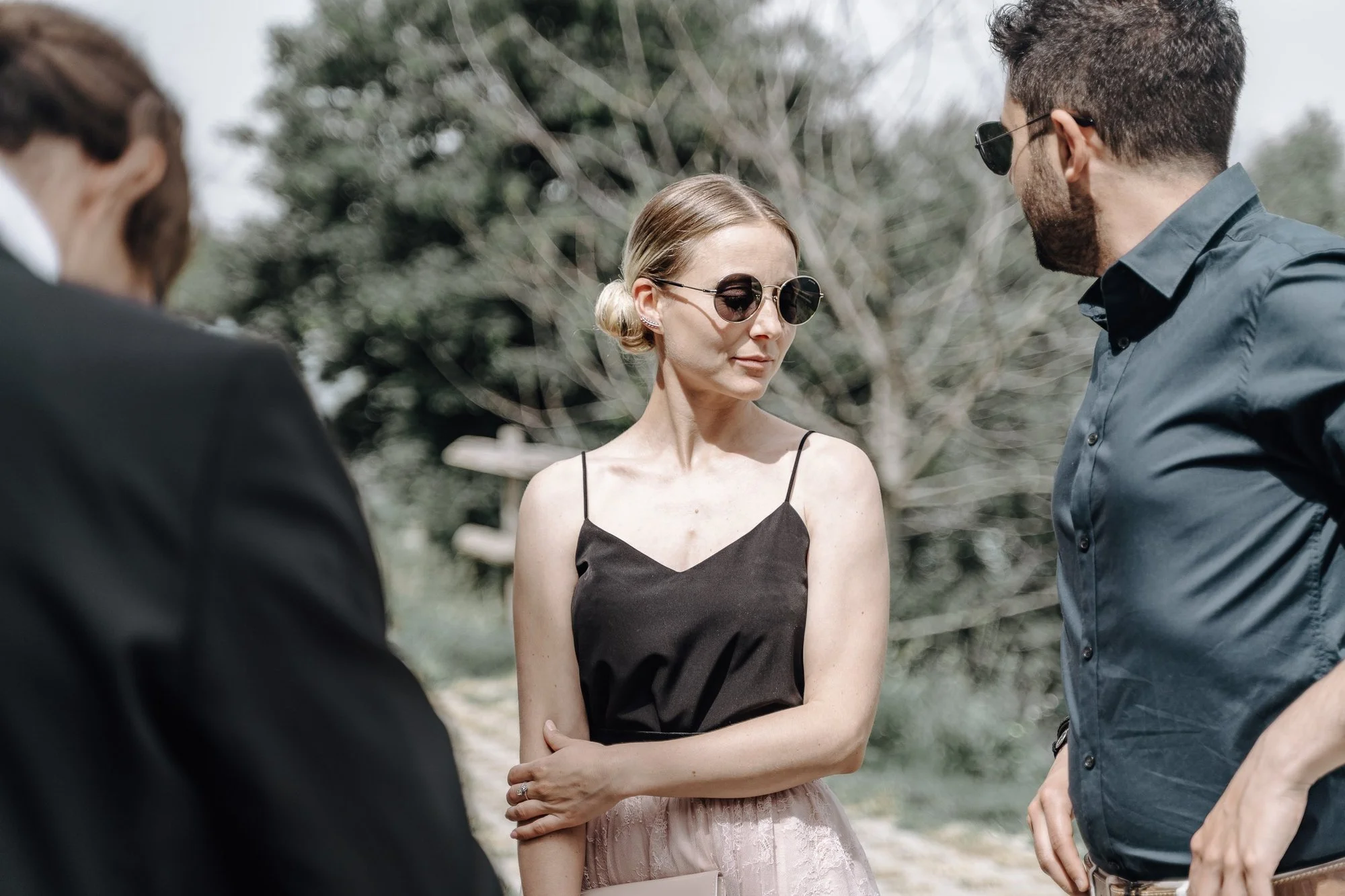 Three people outdoors, two men and one woman, wearing sunglasses. The woman is wearing a black spaghetti strap top and a pink skirt, with her arms crossed. The men are wearing dark shirts, and the man on the right has a beard and short hair. They are