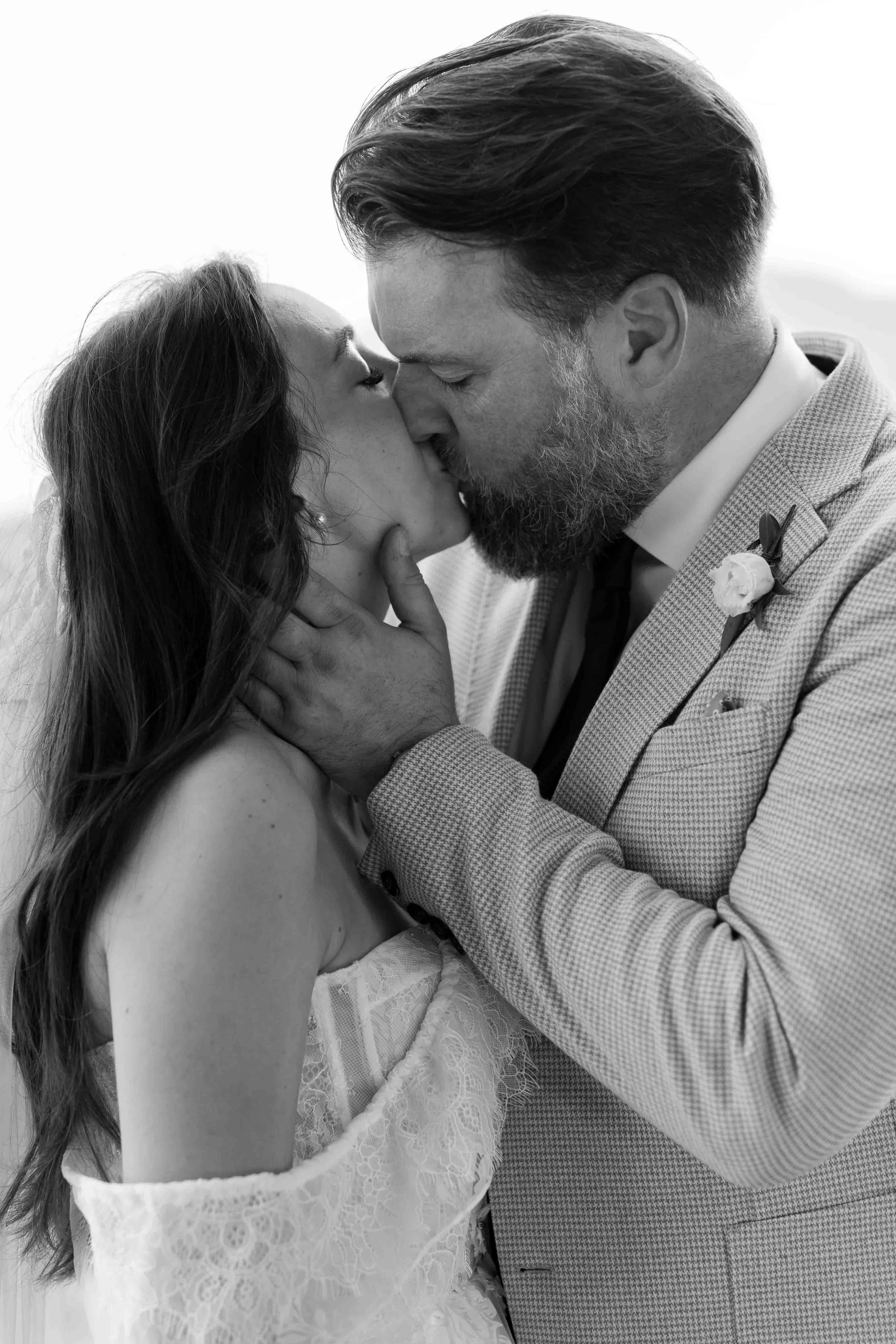 A black and white photo of a man and woman kissing closely, with the man's hand gently touching the woman's face. The woman has long hair and is wearing a lace dress, and the man has a beard, mustache, and is dressed in a checked suit with a boutonni