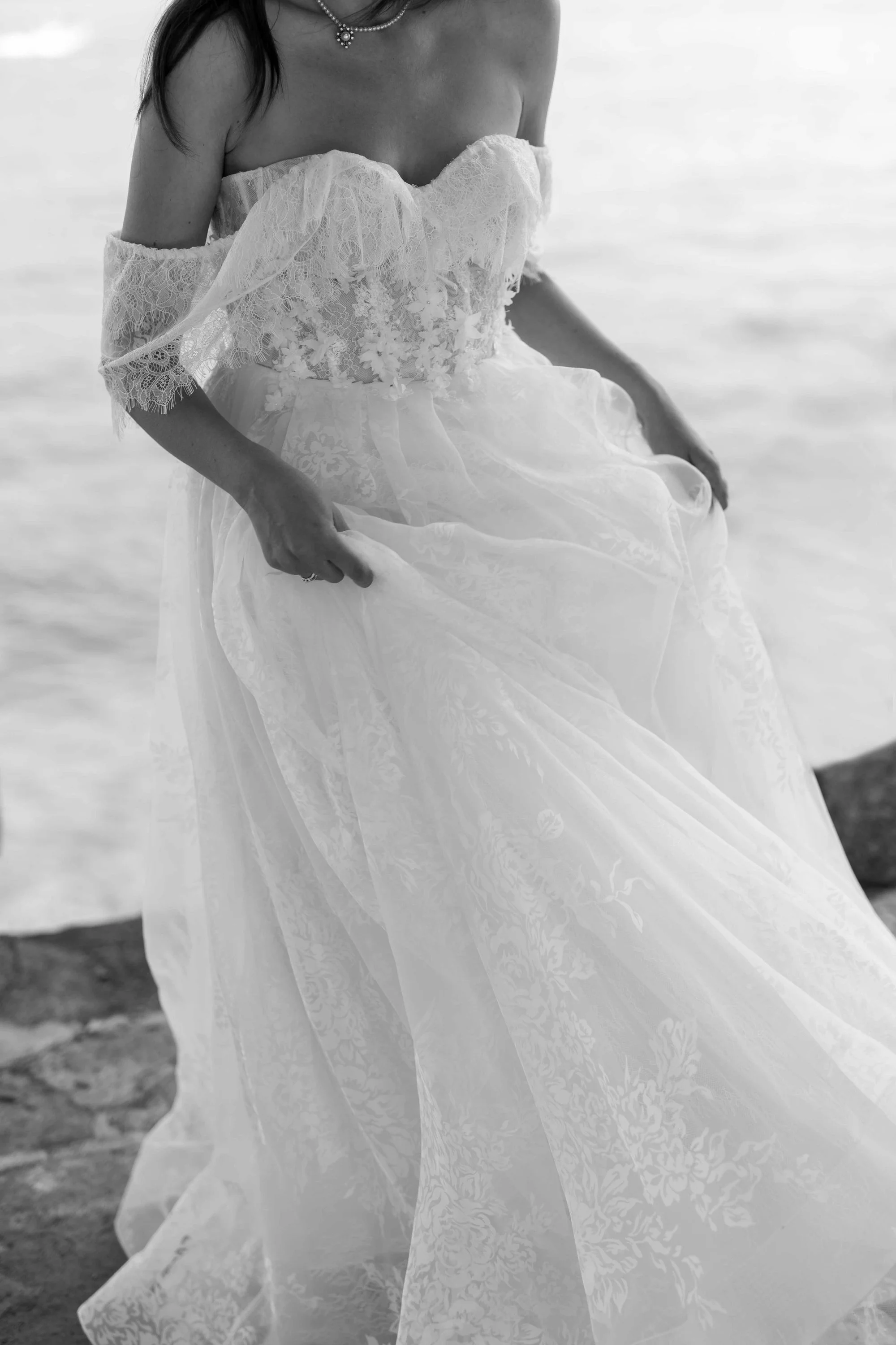 A woman in a wedding dress with off-the-shoulder lace sleeves, sitting near the water, holding the flowing skirt of her dress.