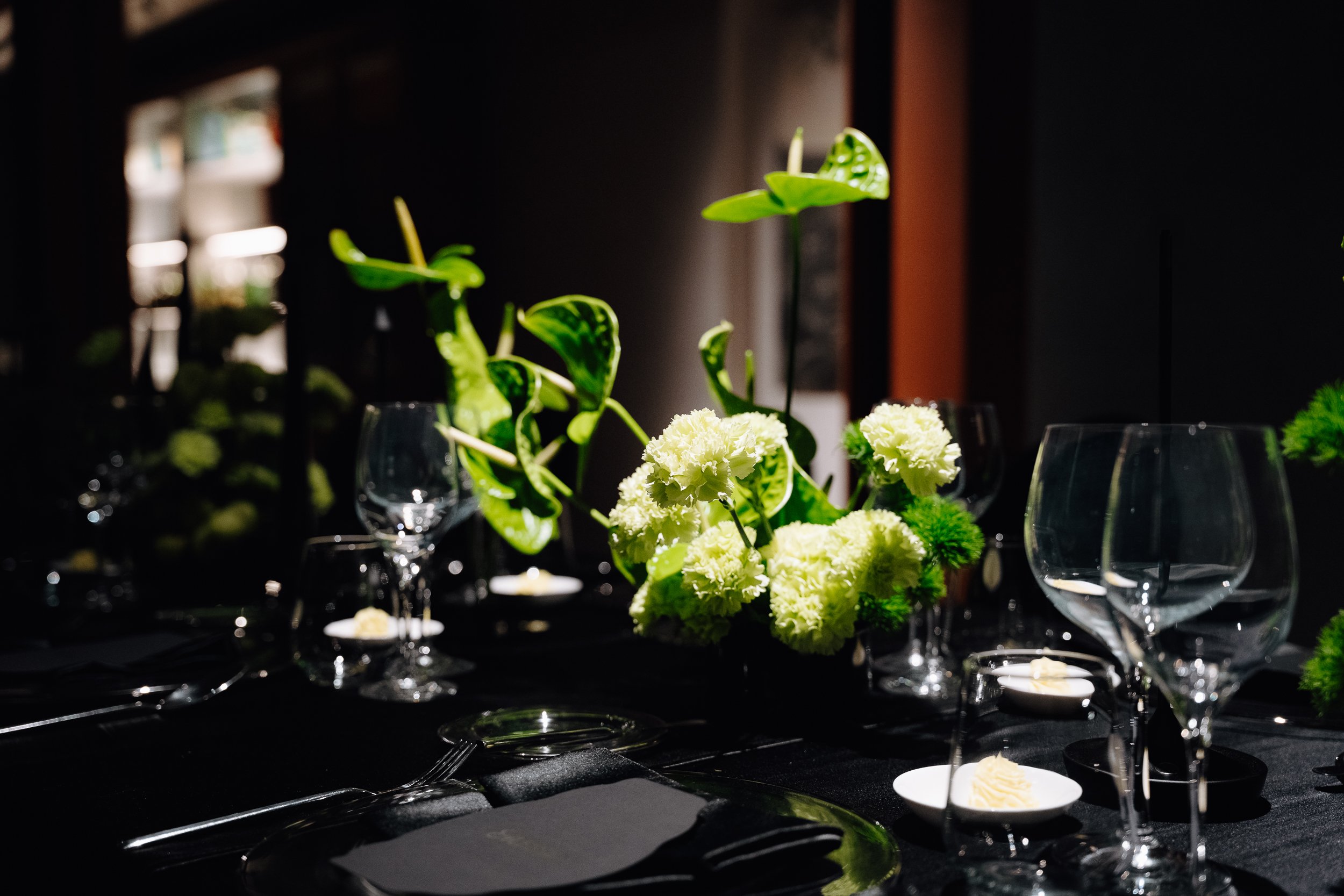 A floral centerpiece with white flowers and green leaves on a dark table, with wine glasses and small white dishes around it, in a dimly lit setting.