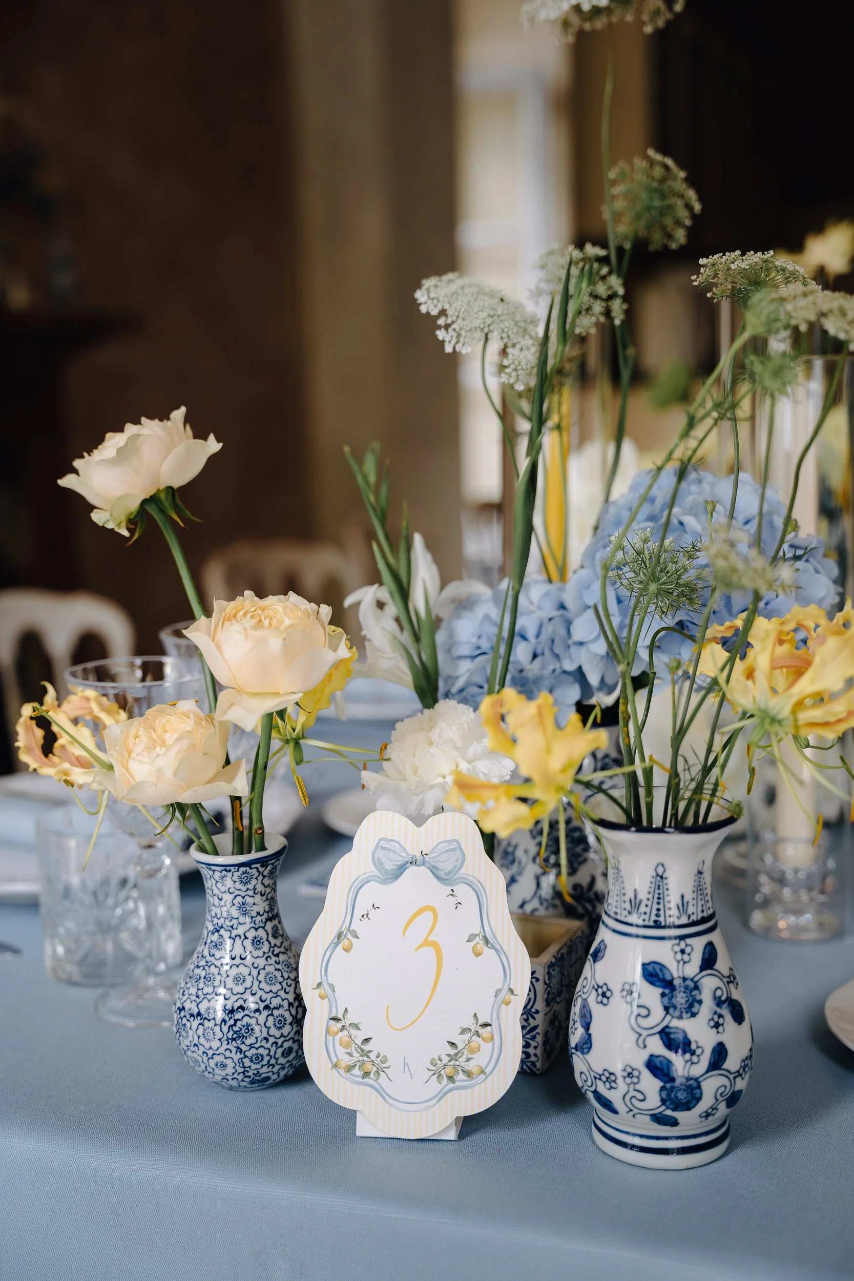 Vases with blue floral patterns holding white, yellow, and light blue flowers, and a table number card with the number 3 in gold, surrounded by a floral design.