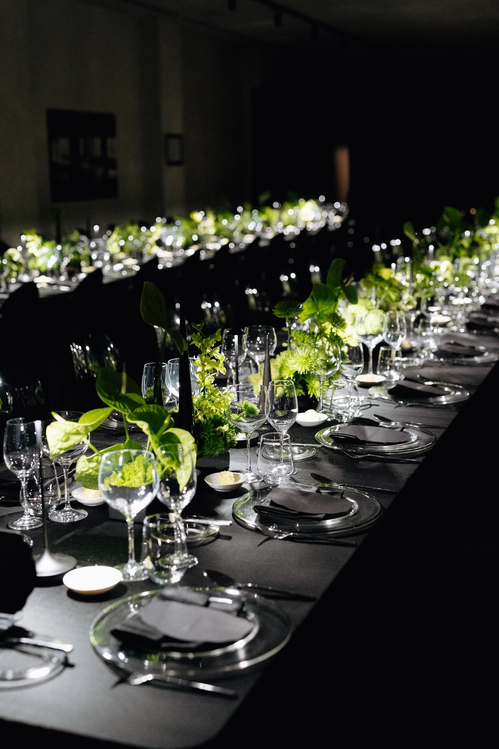 A long dinner table set with wine glasses, plates, napkins, and green floral centerpieces in a dimly lit room, suggesting an elegant event or celebration.