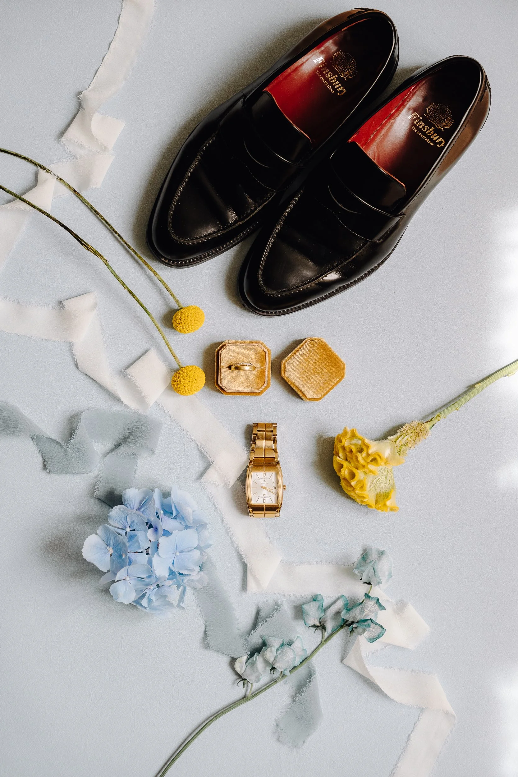 Black leather dress shoes, a gold wristwatch, a ring in a velvet box, and decorative flowers on a light background, with torn white ribbon scattered around.