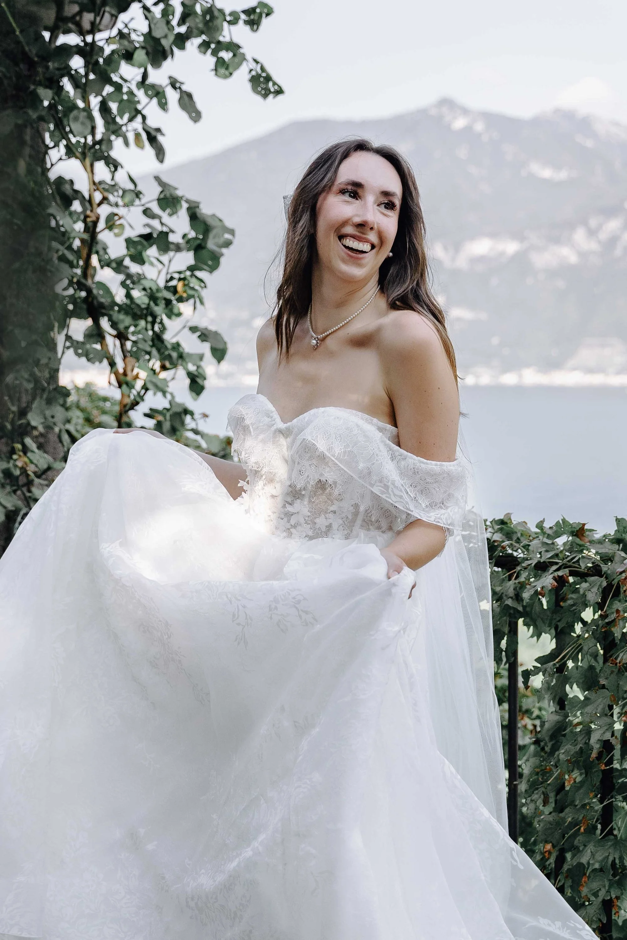 A woman in a white wedding dress smiling outdoors with mountains and a lake in the background.