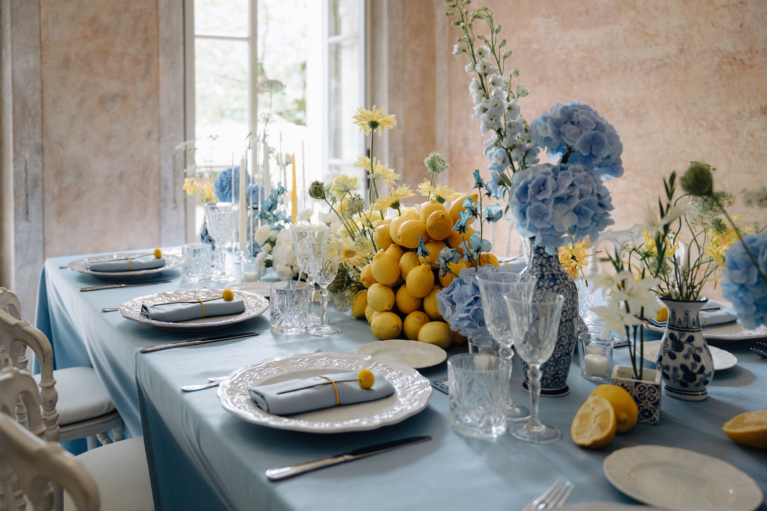 A table set for a meal with a light blue tablecloth, white plates, glassware, silverware, and napkins with yellow rings. The centerpiece includes blue and white flowers, lemons, and other greenery, with a window in the background illuminating the roo