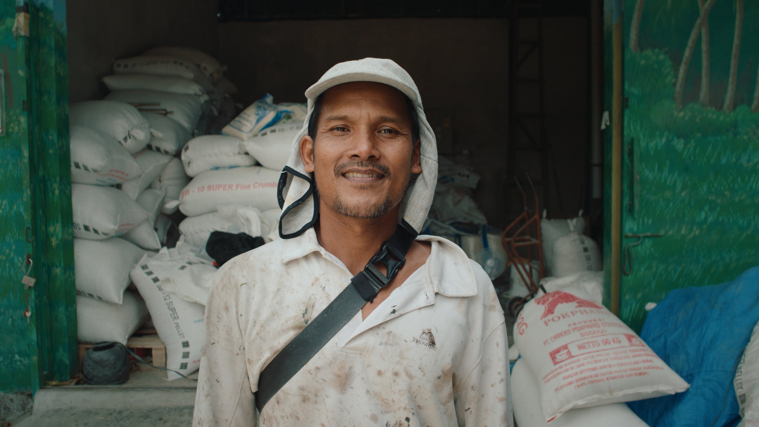 Smiling man wearing a light-colored shirt and a wide-brimmed hat standing outside a storage area filled with sacks.