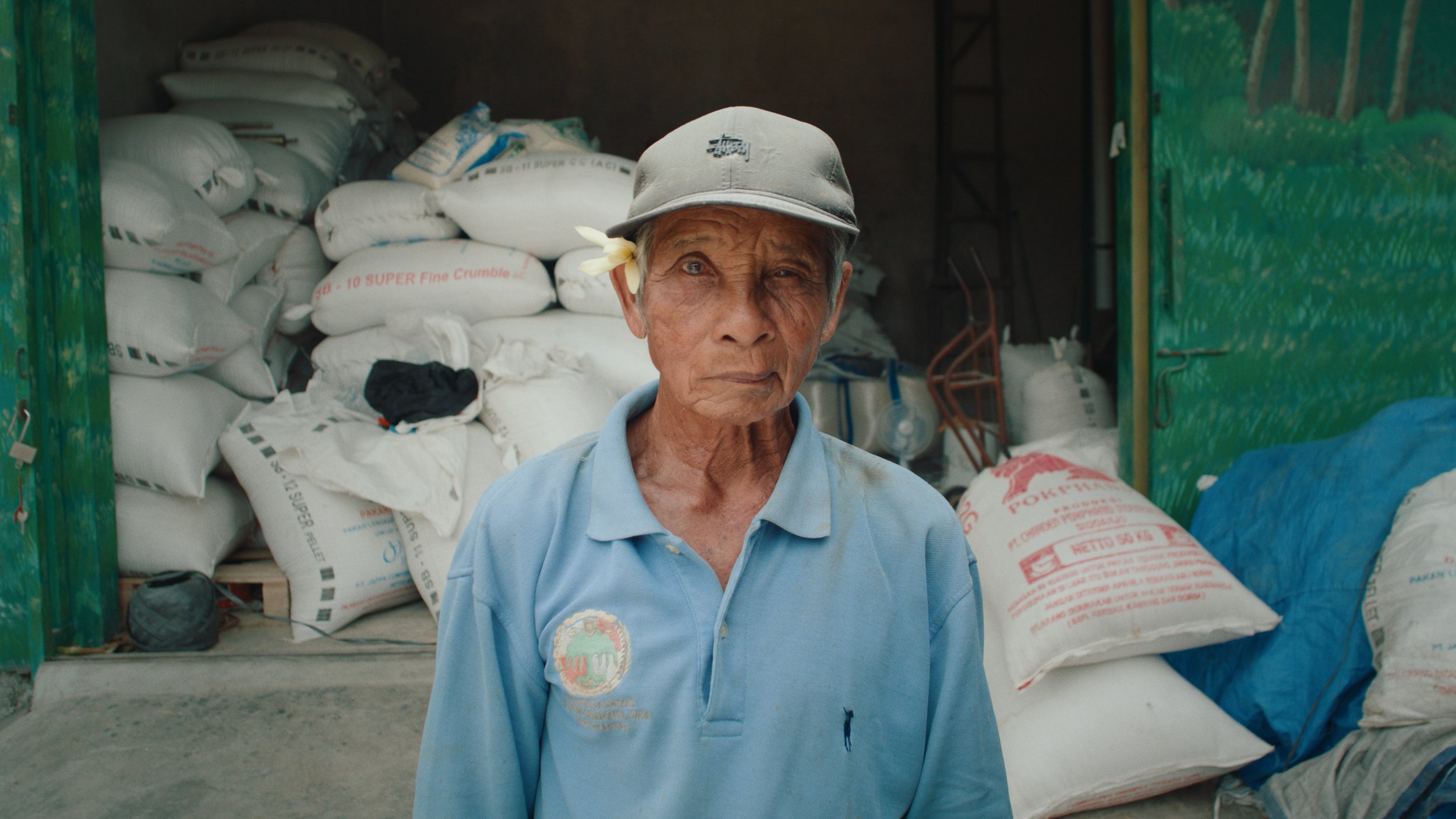 An elderly woman wearing a gray cap and a light blue polo shirt, standing in front of large sacks of rice or flour stacked inside a warehouse. The woman has a flower tucked behind her ear and a serious expression on her face.
