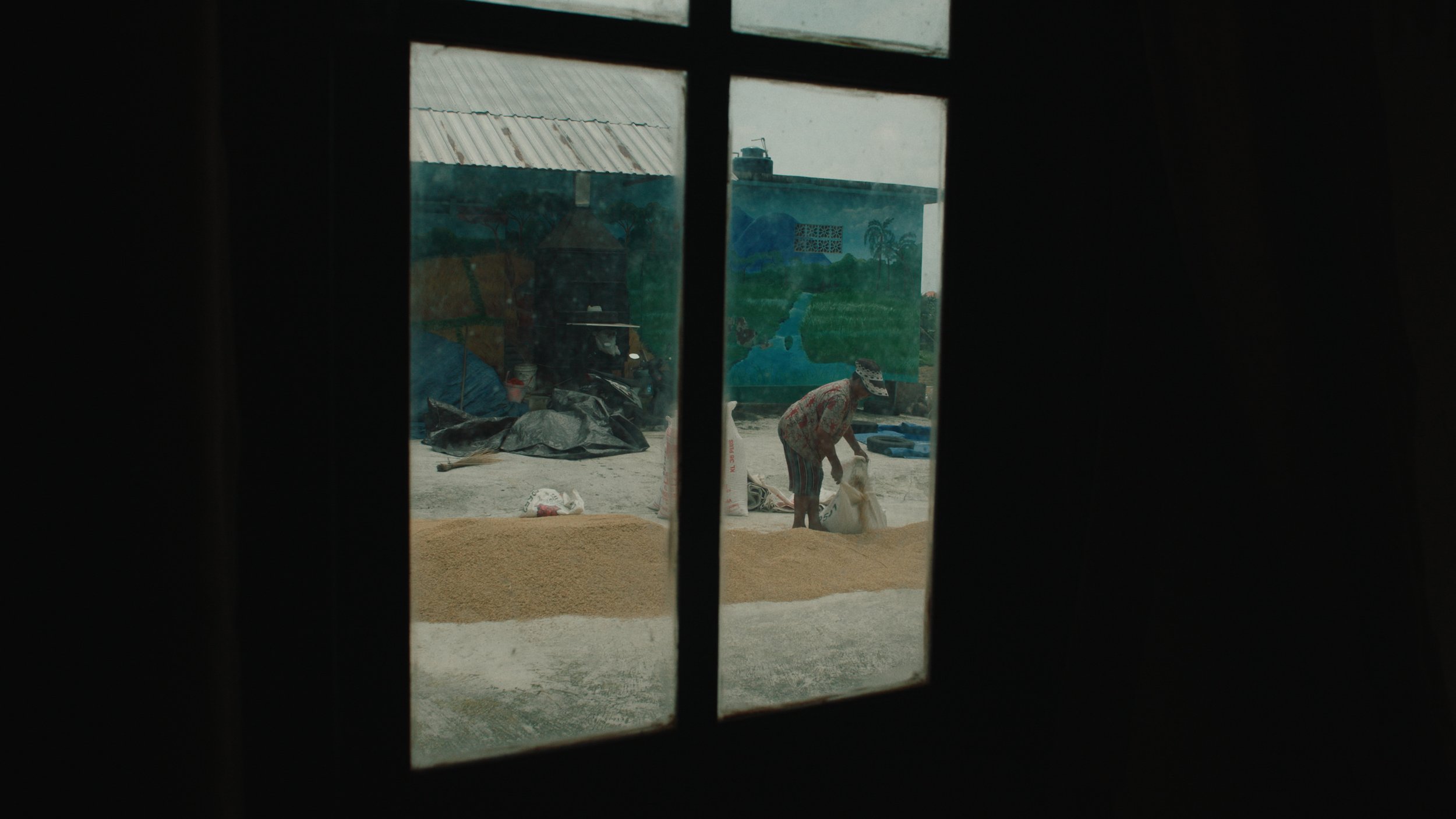 A person is working outdoors, seen through a window with black framing, with a mural of a landscape in the background.