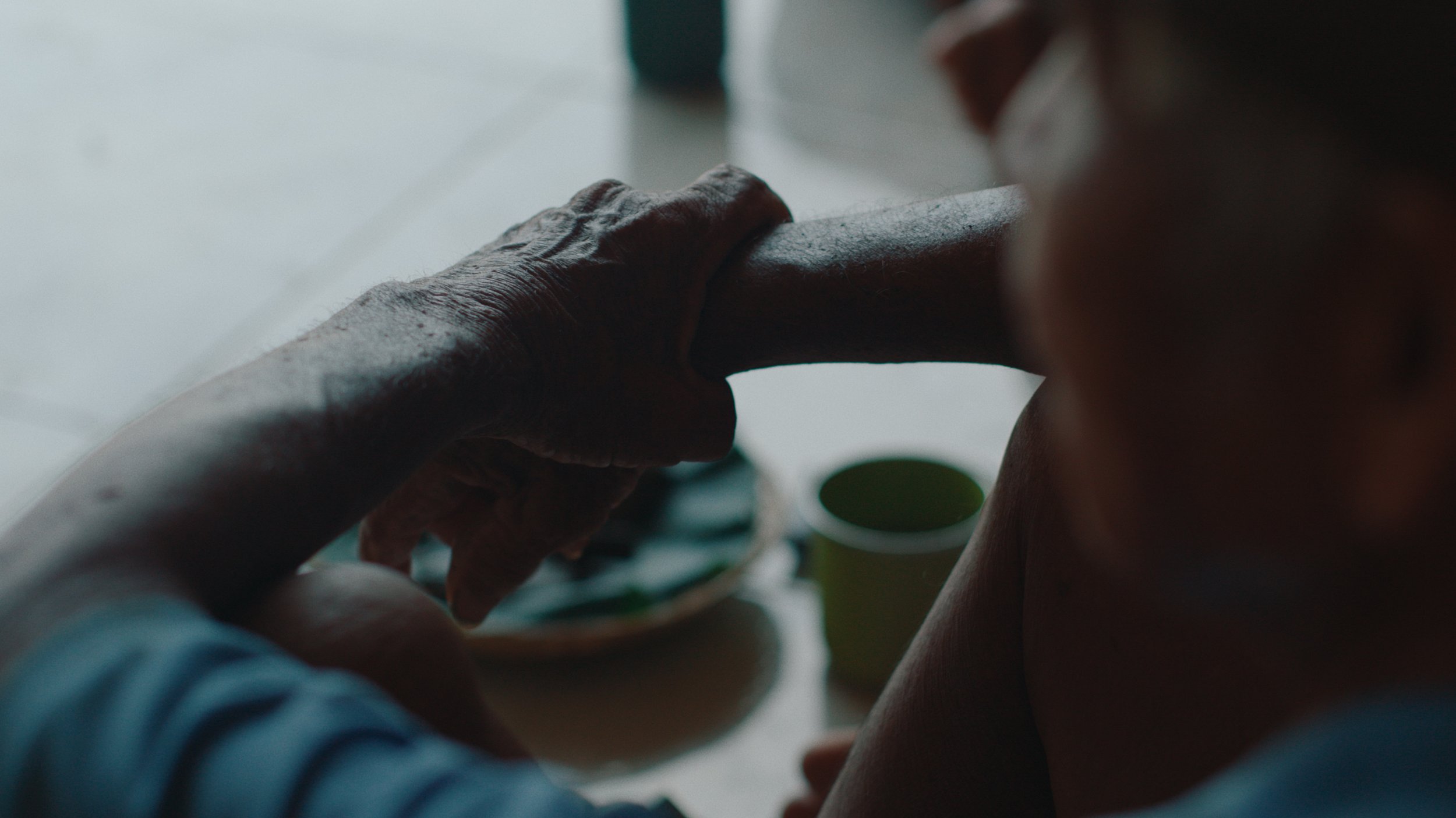 A person is feeding a baby with a spoon while sitting at a table, with a bowl and cup nearby.