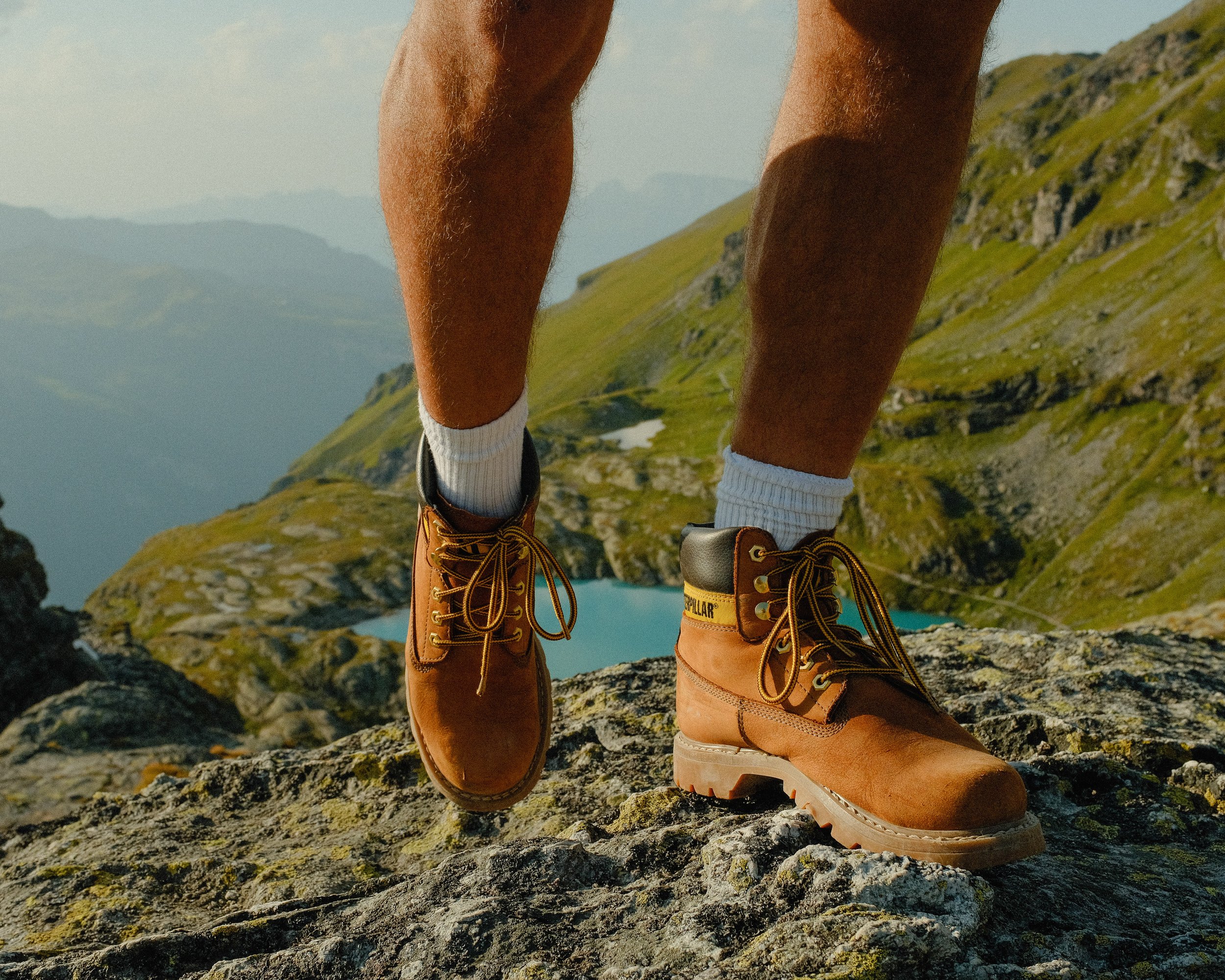 A person wearing tan work boots and white socks standing on a rocky mountain terrain with a scenic view of a lake and hills in the background.
