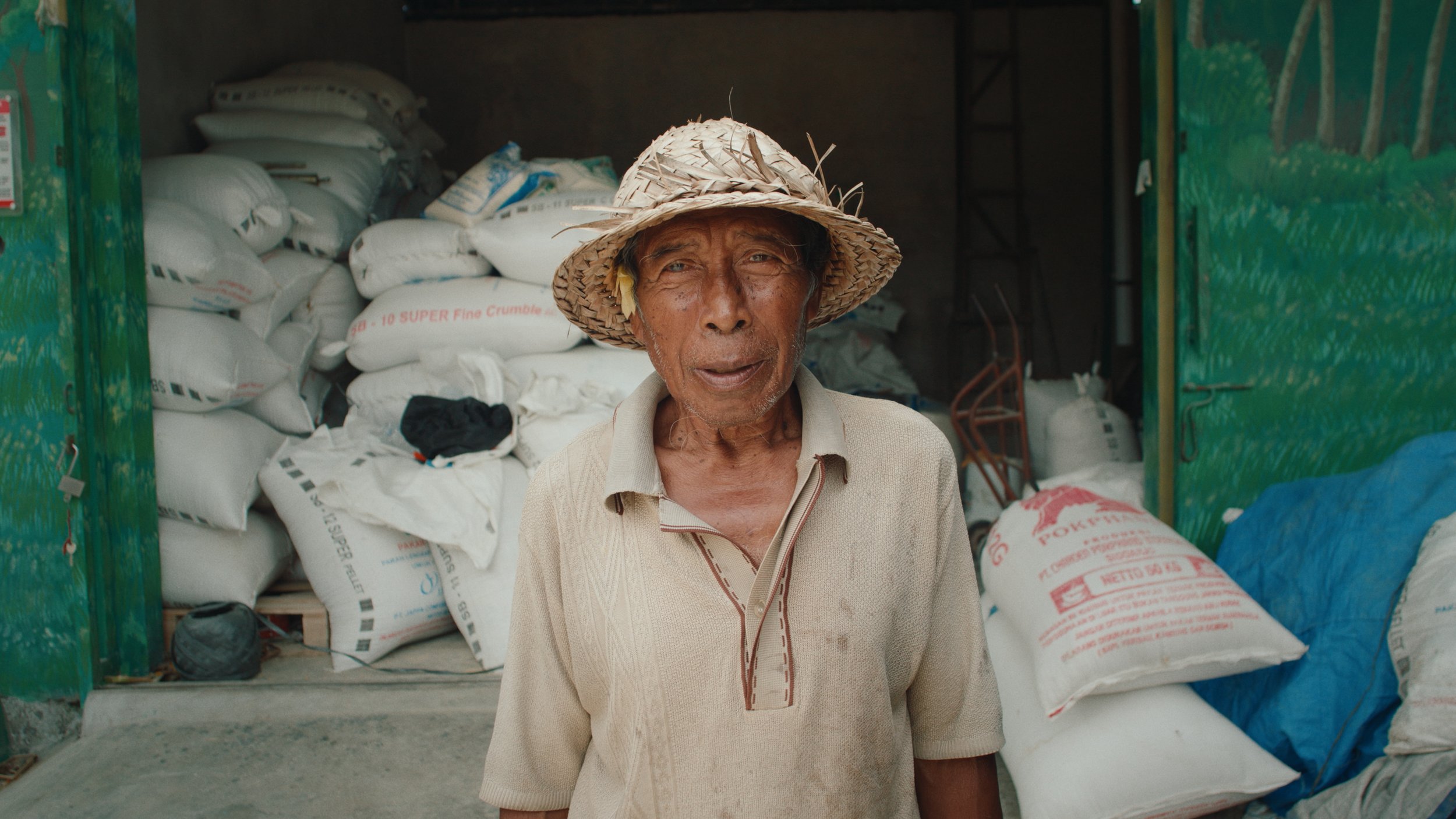 An elderly man in a straw hat standing in front of large bags of rice or grain inside a storage area or shop.