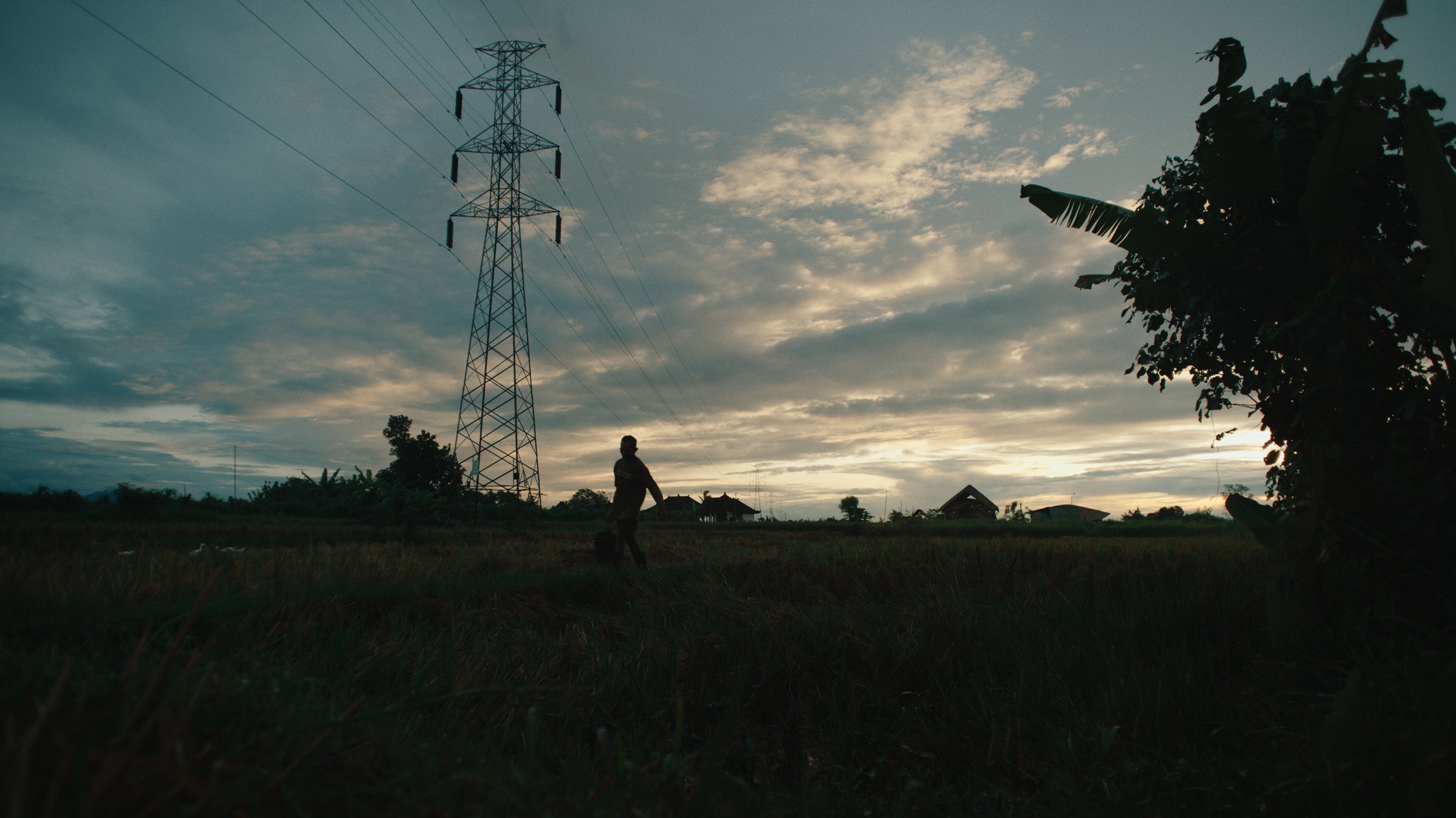 Silhouette of a person walking in a field during sunset with an electrical tower and scattered houses in the background.