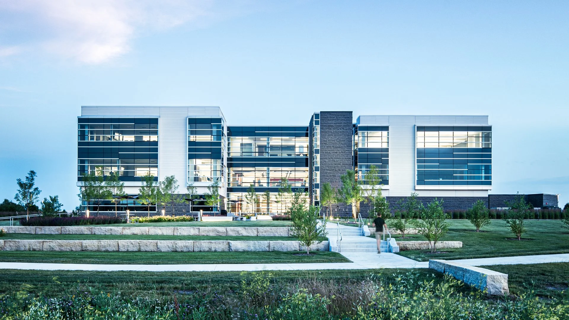 Modern multi-story office building with glass windows, surrounded by landscaped lawn and trees, under a clear sky. IMT Insurance Building in West Des Moines, IA.