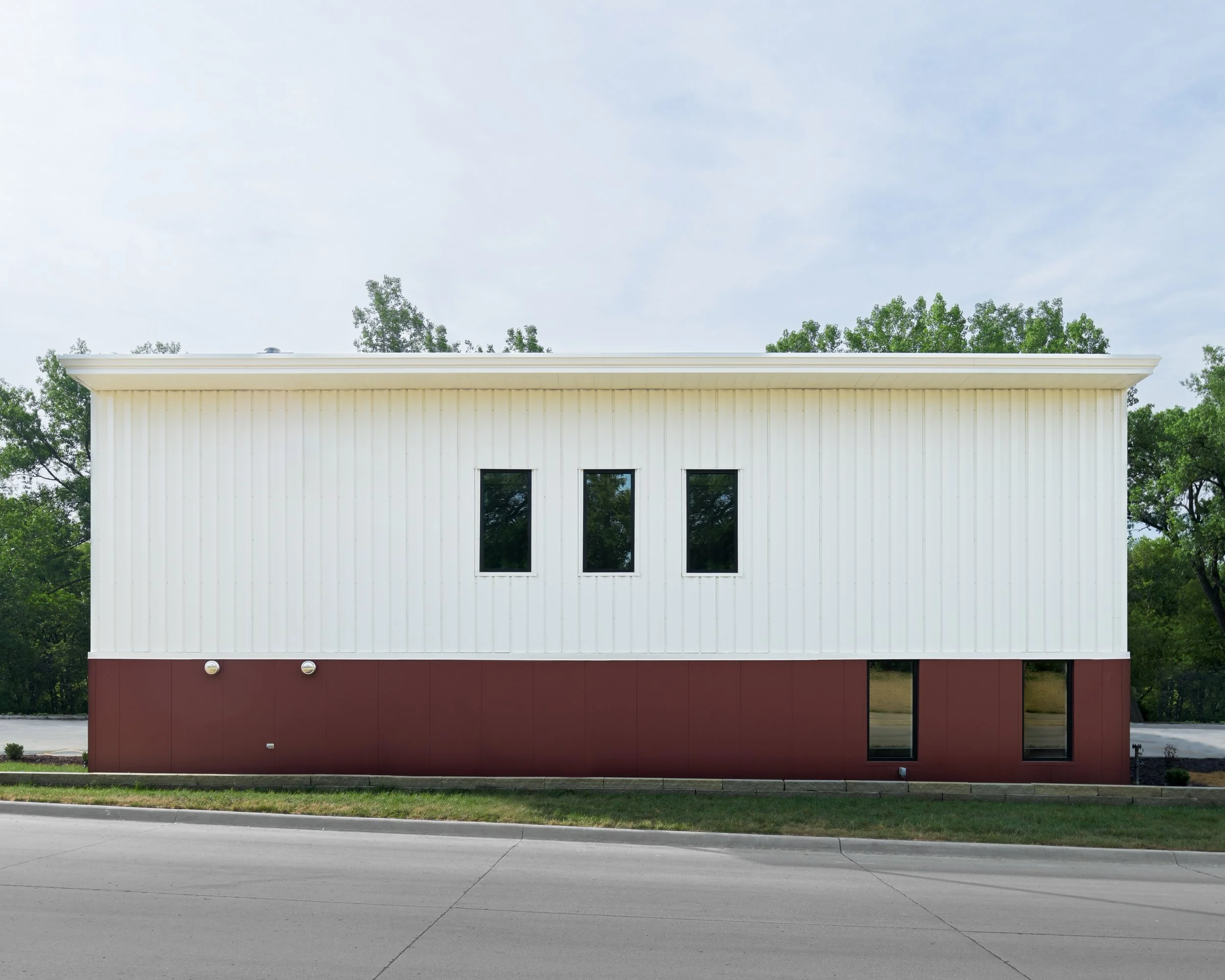 Photo of a modern white building with dark red base, three windows visible, and a flat roof, surrounded by greenery and a cloudy sky.