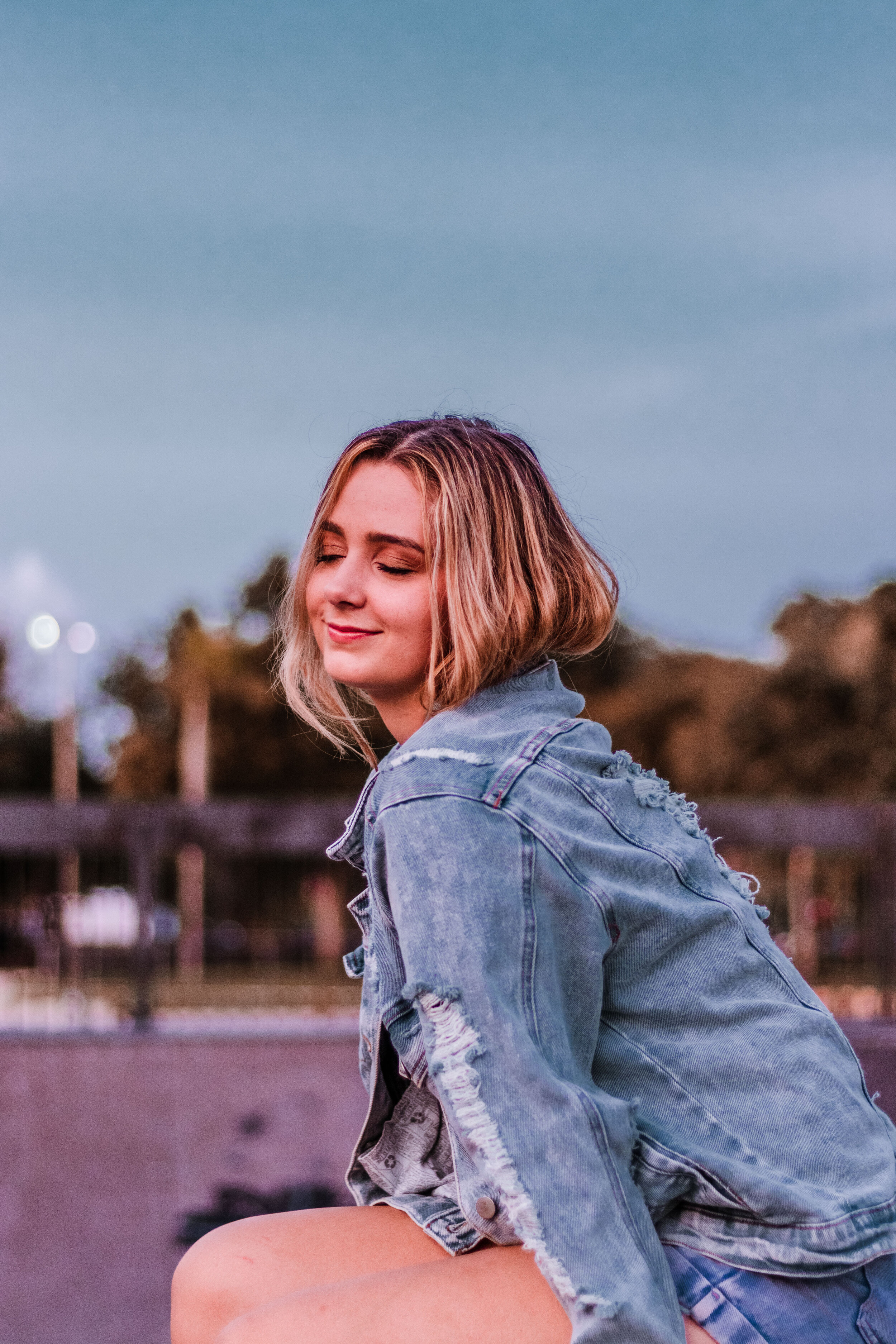 Young woman with blonde hair wearing a distressed denim jacket, sitting outdoors with her eyes closed and smiling gently during the evening.